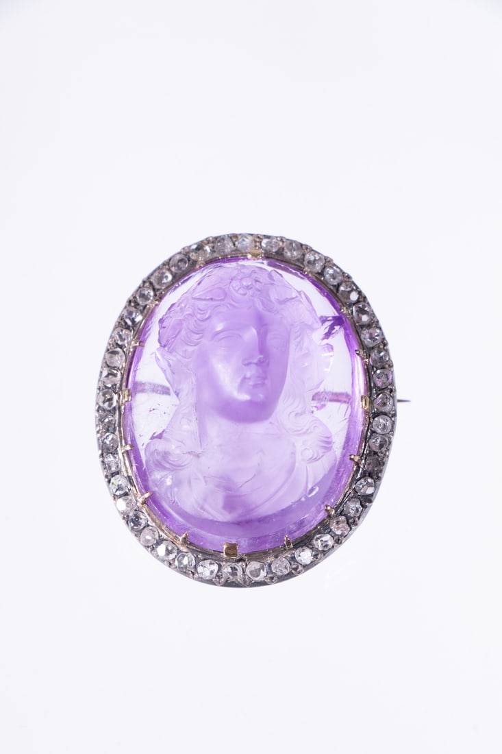 RARE AMETHYST CAMEO BROOCH WITH DIAMONDS, 19TH CENTURY: An oval amethyst cameo brooch, late 1800s, finely carved in high relief with a neoclassical portrait of a woman. The cameo is set in a 14K yellow gold mounting with a prong setting and a pin mechanism