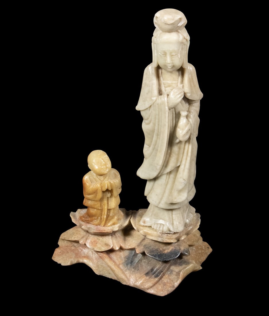 CHINESE CARVED SOAPSTONE FIGURE OF GUANYIN, EARLY 20TH CENTURY: CHINESE CARVED SOAPSTONE FIGURE OF GUANYIN, EARLY 20TH CENTURY A carved soapstone figure of Guanyin holding a vase, accompanied by a kneeling acolyte on a rocky base. The stone exhibits light