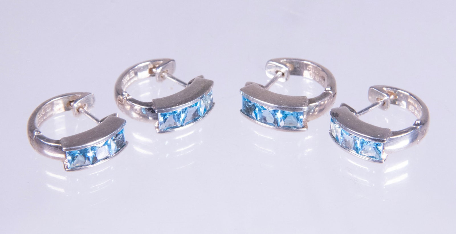 (2) PRS STERLING SILVER AND BLUE TOPAZ HOOP EARRINGS: (2) PRS STERLING SILVER AND BLUE TOPAZ HOOP EARRINGS A group of two pairs of sterling silver hoop earrings, each set with (3) princess-cut blue topaz stones measuring approximately 4.09 mm. The stones