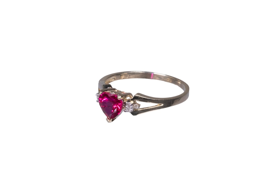 10K GOLD SYNTHETIC RUBY AND DIAMOND RING: 10K GOLD SYNTHETIC RUBY AND DIAMOND RING The ring set with a heart-shaped synthetic ruby, flanked on each side by (3) diamond melee accents, for a total of (6) diamonds. Mounted in a split-shank 10K