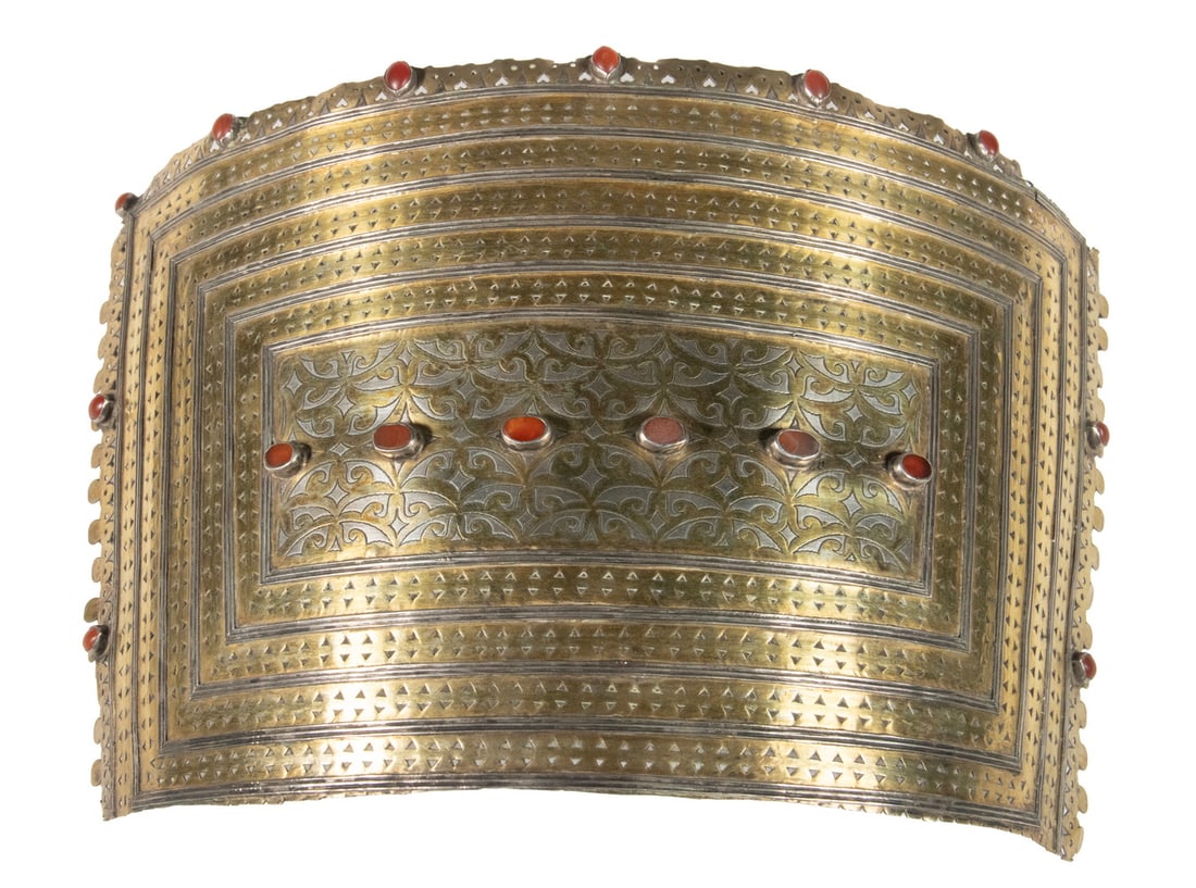 TEKKE TURKOMAN GILT SILVER AND CARNELIAN EGME HOOD ORNAMENT: TEKKE TURKOMAN GILT SILVER AND CARNELIAN EGME HOOD ORNAMENT A curved silver and gilt egme hood ornament of traditional Tekke Turkmen form. The surface is decorated with punched geometric motifs and