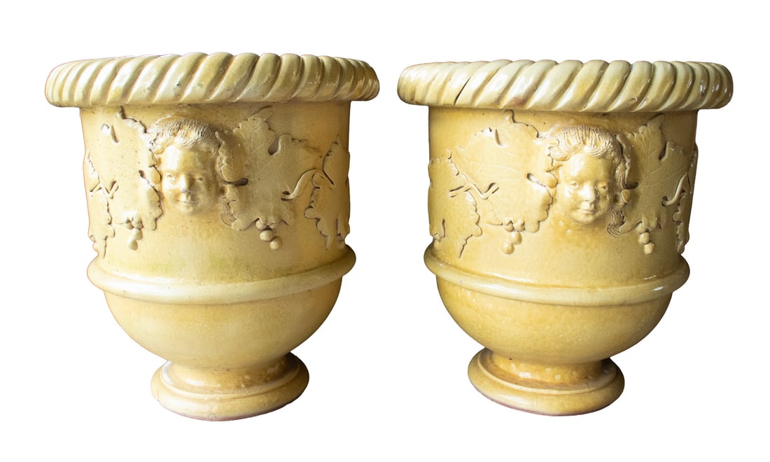 PAIR OF GARGANTUAN ROMANESQUE YELLOW GARDEN URNS: PAIR OF GARGANTUAN ROMANESQUE YELLOW GARDEN URNS Massive Terra Cotta Exterior Urns in butter yellow heavy glaze, featuring cupid heads. Fine condition. OS, Each: 29 x 24 x 24 in. (73.7 x 61 x 61 cm.)