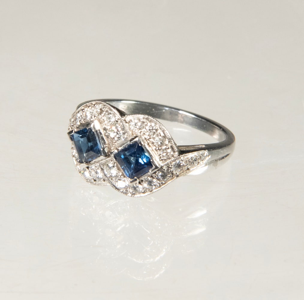 ART DECO PLATINUM, DIAMOND, AND SAPPHIRE RING: ART DECO PLATINUM, DIAMOND, AND SAPPHIRE RING An Art Deco platinum ring set with (2) square step-cut blue sapphires, each measuring approximately 4.05 mm, framed by (24) round diamonds in a fine