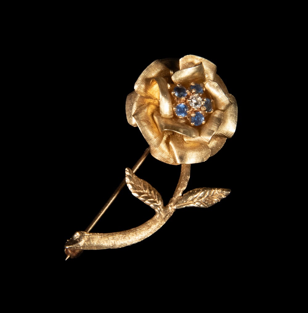 14K GOLD, DIAMOND, AND SAPPHIRE ROSE BROOCH: 14K GOLD, DIAMOND, AND SAPPHIRE ROSE BROOCH A finely textured 14K yellow gold brooch designed as a blooming rose with scrolling stem and foliate leaves. The flower head is centered by a small round