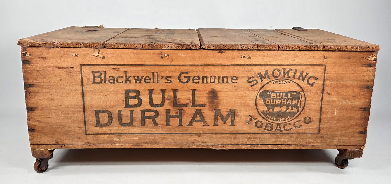 BLACKWELL'S GENUINE BULL DURHAM SMOKING TOBACCO WOOD BOX: REMAINS IN FAIR ORIGINAL CONDITION, SHOWING WEAR. LID HAS BEEN ADDED AND CASTOR WHEELS APPLIED.