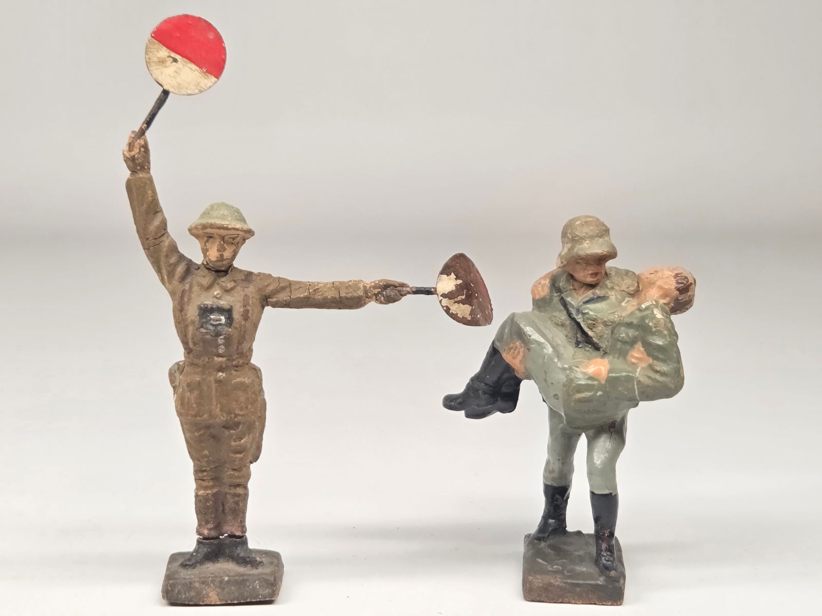 GERMAN ELASTOLIN SIGNAL & SOLDIER CARRYING WOUNDED: BOTH REMAIN IN FAIR CONDITION. SIGNALER SHOWING BREAK AT FEET AND MULTIPLE CRACKS. ONE OF THE SIGNALS IS BENT. THE SOLDIERS CARRYING WOUNDED SOLDIER SHOWING WEAR.