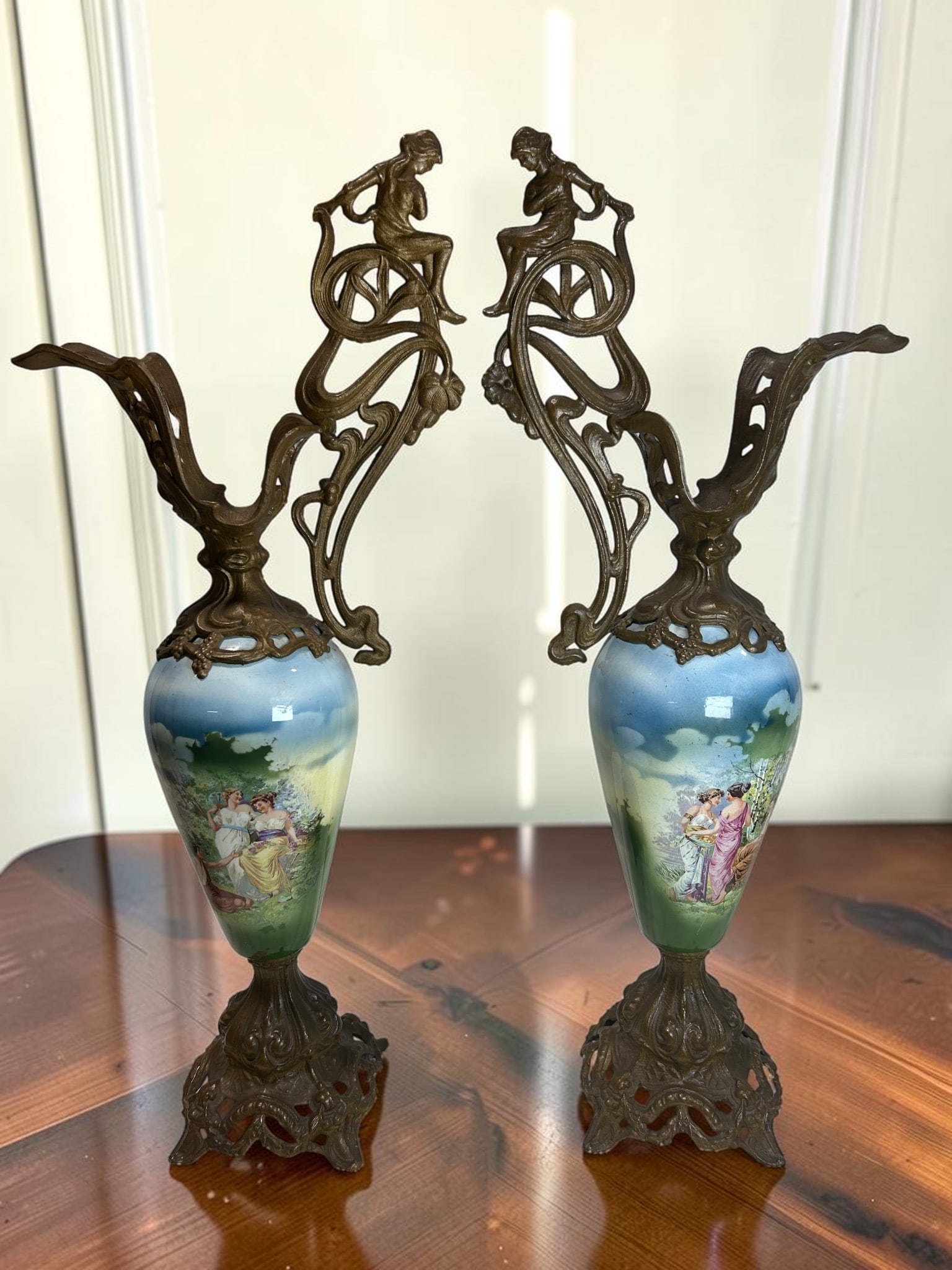 Pair of Antique Art Nouveau Figural Bronze & Hand-Painted Porcelain Ewers: This exquisite pair of antique Art Nouveau figural ewers features elegant hand-painted porcelain bodies mounted in ornate flowing bronze frames with sculptural female figures perched at the handles. R