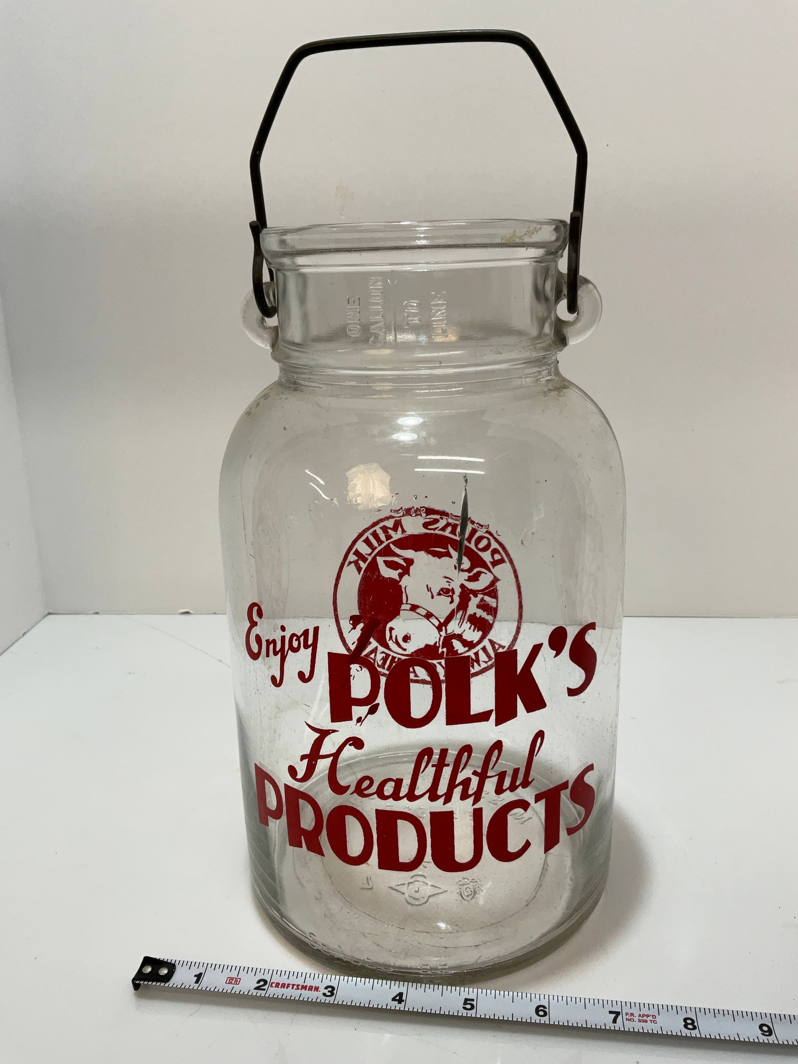 Glass Polk's Milk Jug One Gallon: One gallon Polk's Milk Jug. "Enjoy Polk's Healthful Products" is on one side of the jug, and "Polk's Milk Always Ahead" cow logo on the other side of the jug. Metal handle. There is a crack in the gla