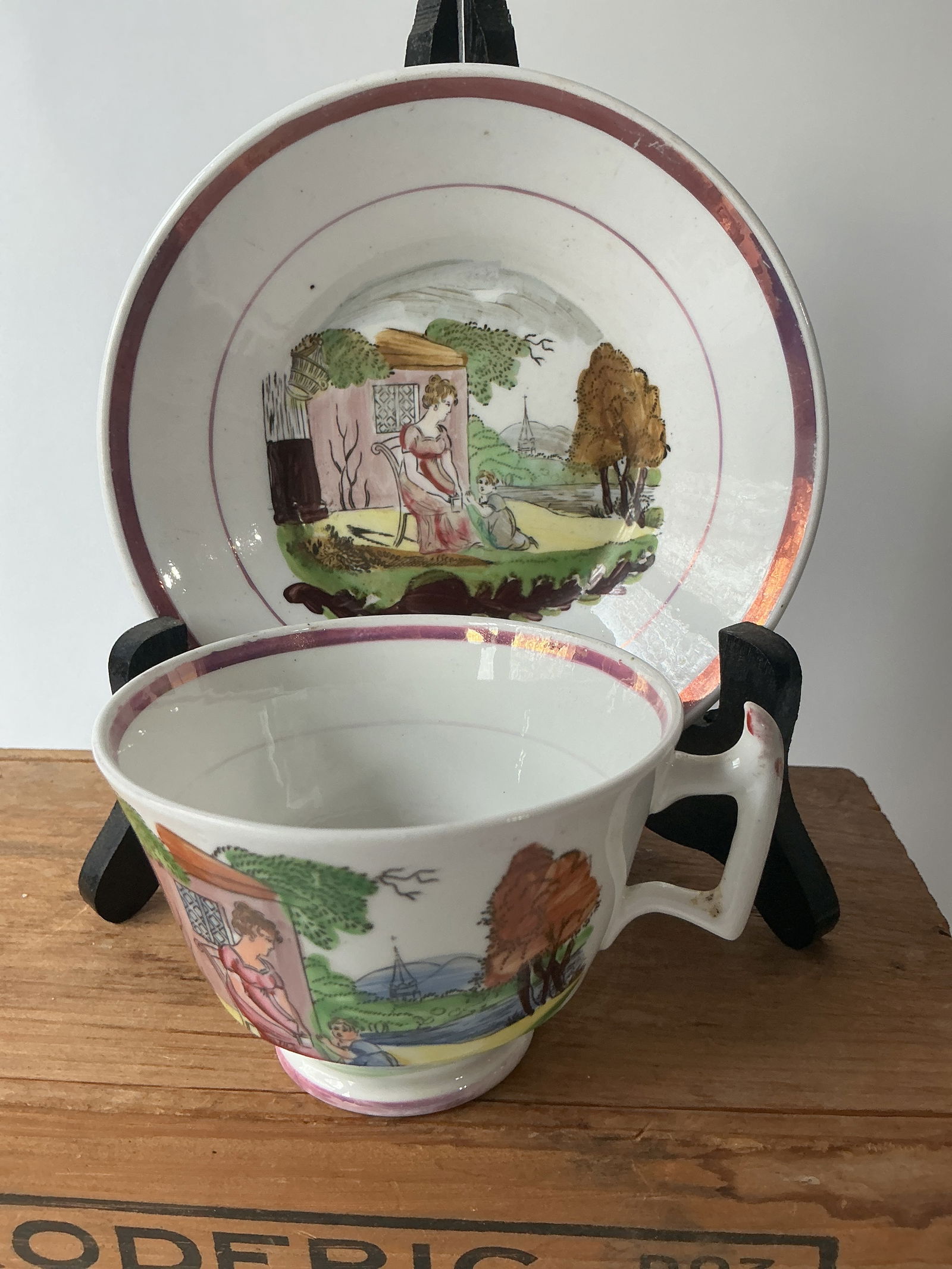 Early Cup and Saucer: Ca/ 1840s English bone china handled cup and saucer with two outlined prints of domestic scenes heavily embelished with fired on over-paints. Cup and saucer have pink lustre added and are in very good