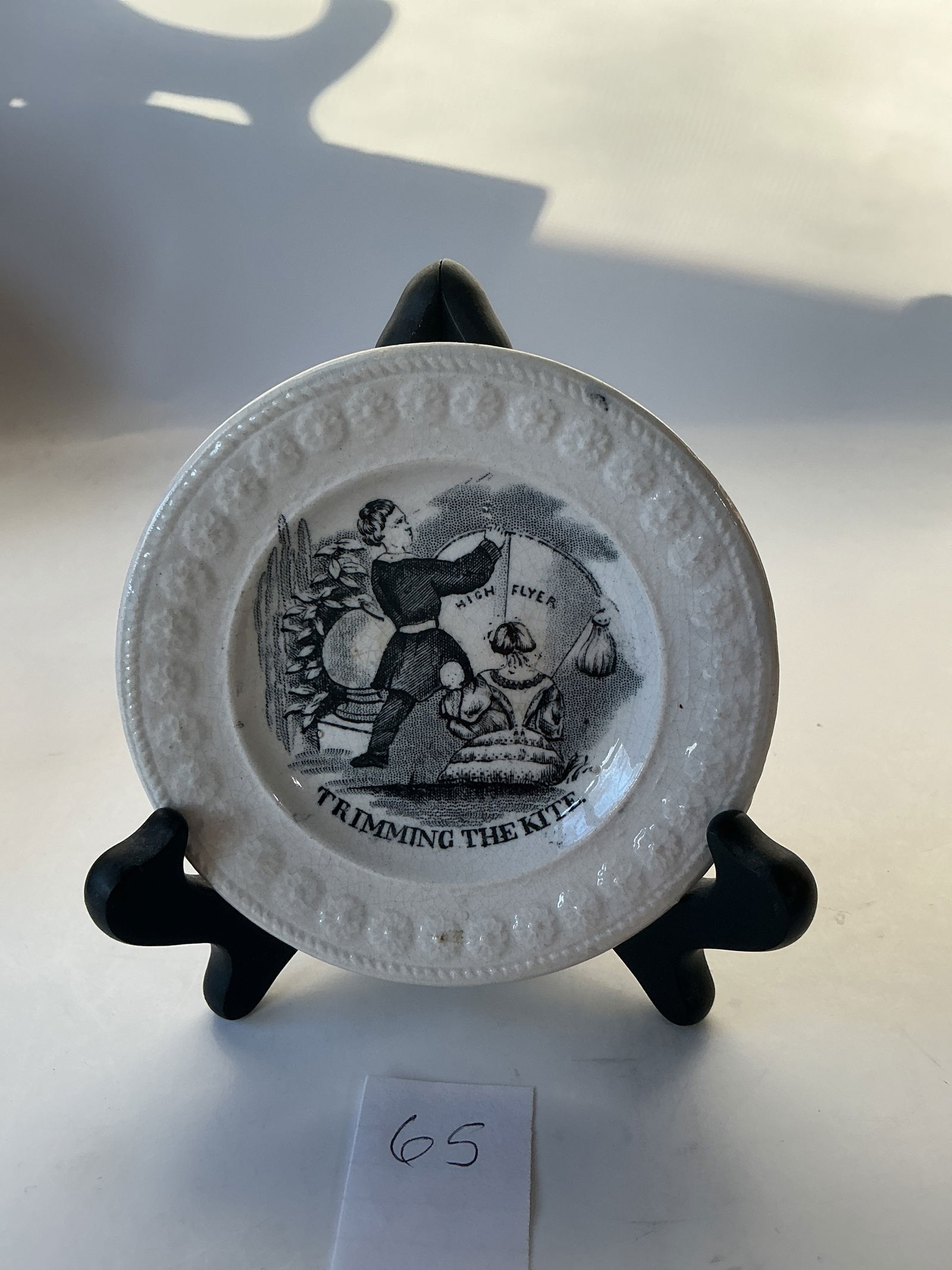 Miniature Pastime Plate: This is a ca. 1850s plate titled "Trimming The Kite". It is 4 1/4" wide and has a floral embossed rim. The girl in front is holding a doll. There are two tiny chips visible on the underside.