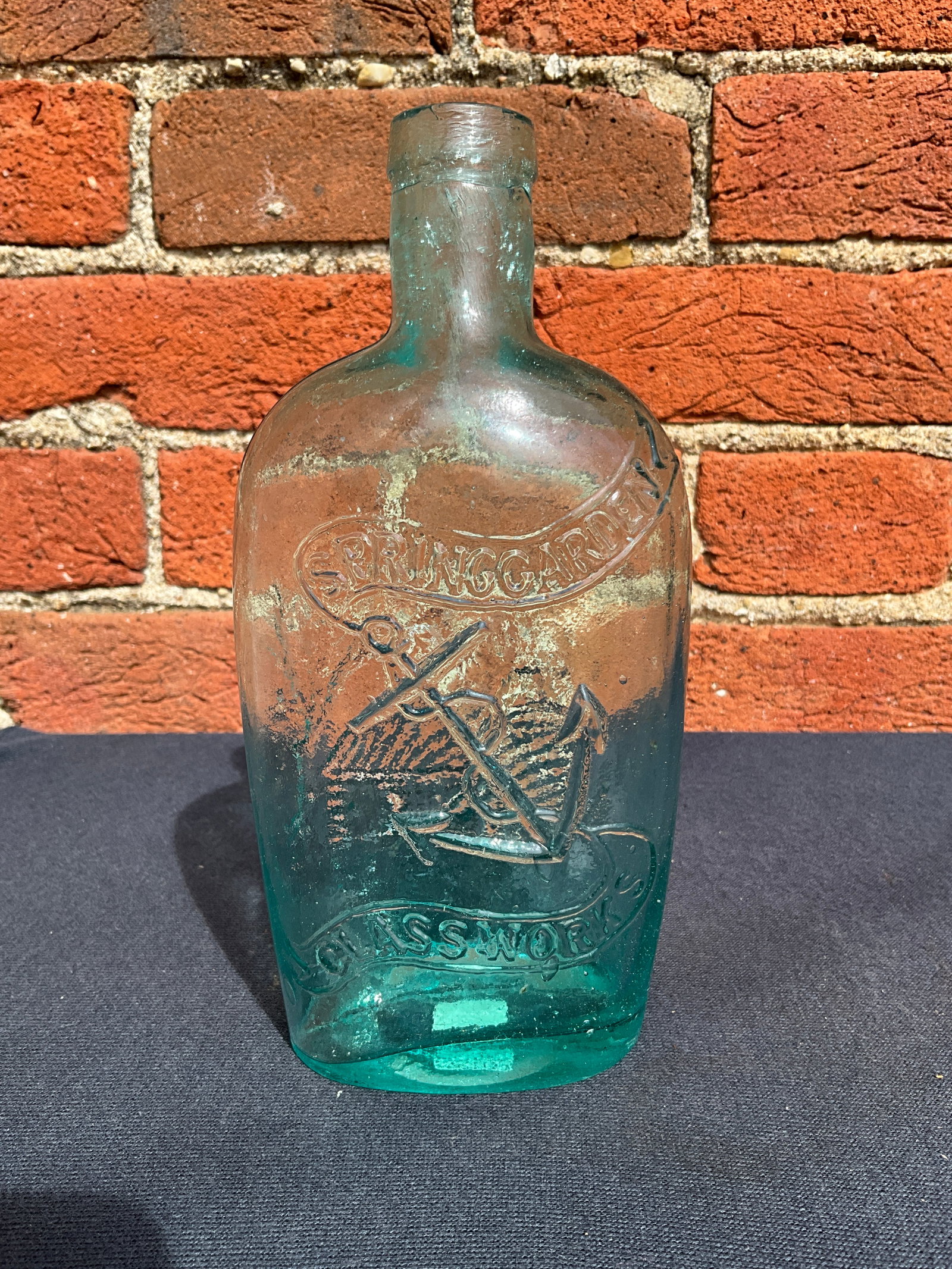 Historical Flask Embossed Spring Garden Glass Works W/ an Anchor on Front, and a cabin on Reverse: Here is an antique historical flask embossed spring garden glass works with an anchor on one side, and a cabin scene on the opposing side. Has a very crudely applied ring lip. No cracks, chips, dings.
