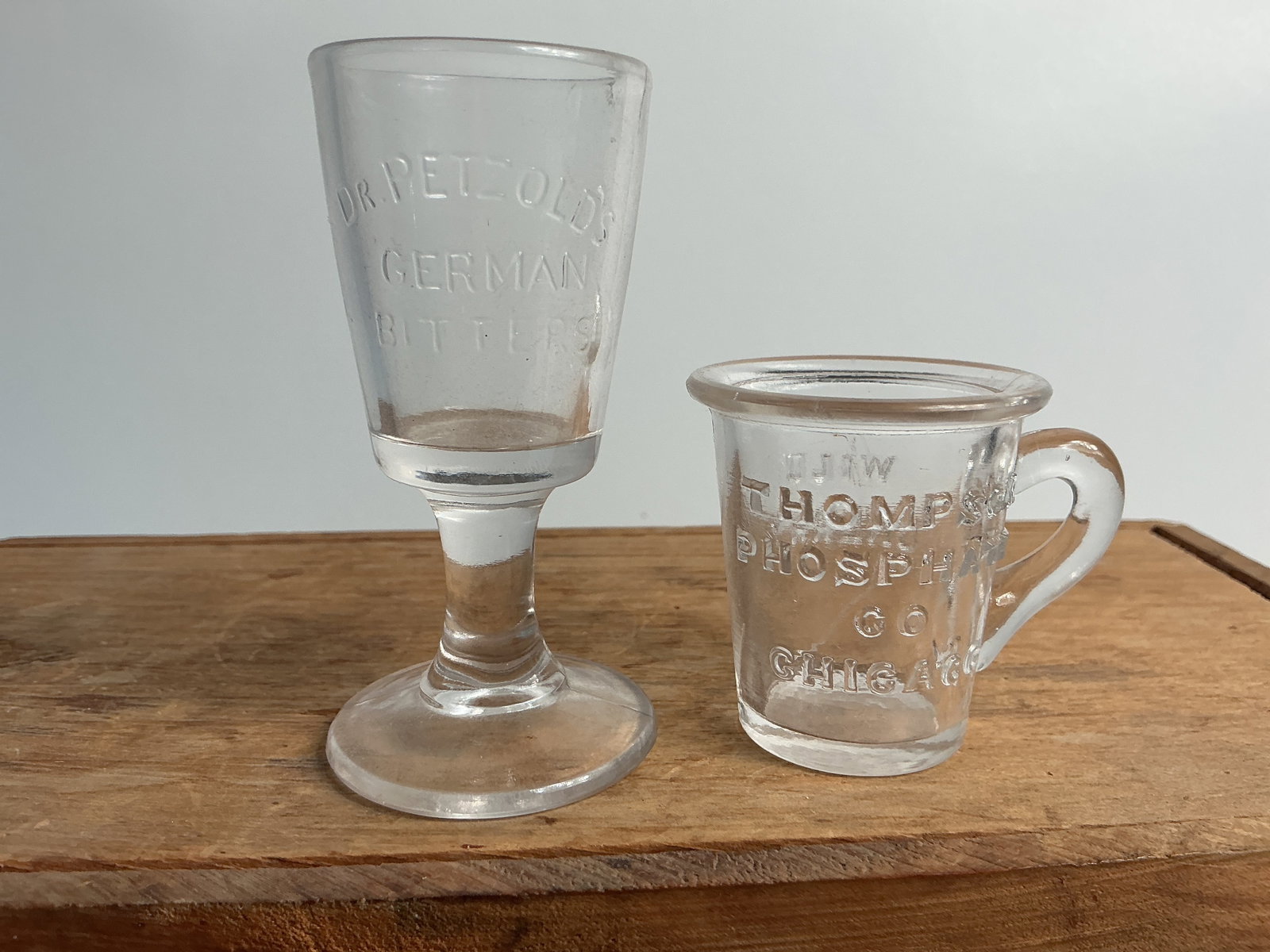2 Embossed Dose Glasses - Dr. Petzold's German Bitters & Thomson Phosphate Co.: Glass one is a Dr. Petzold's German Bitters, clear, 3 1/4" tall. Glass two is Thompson Phosphate co. Chicago Wild Cherry, clear, 2" tall. Both are in great condition.