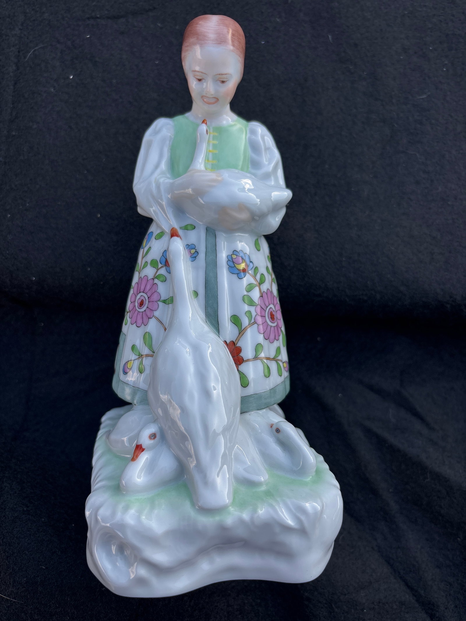 Vintage Herend Hungary 8 inch statue girl with goose: Figure depicting a peasant girl holding a goose while several geese gather at her feet. The girl is dressed in traditional attire, featuring a long pleated skirt with floral apron. Each element is met
