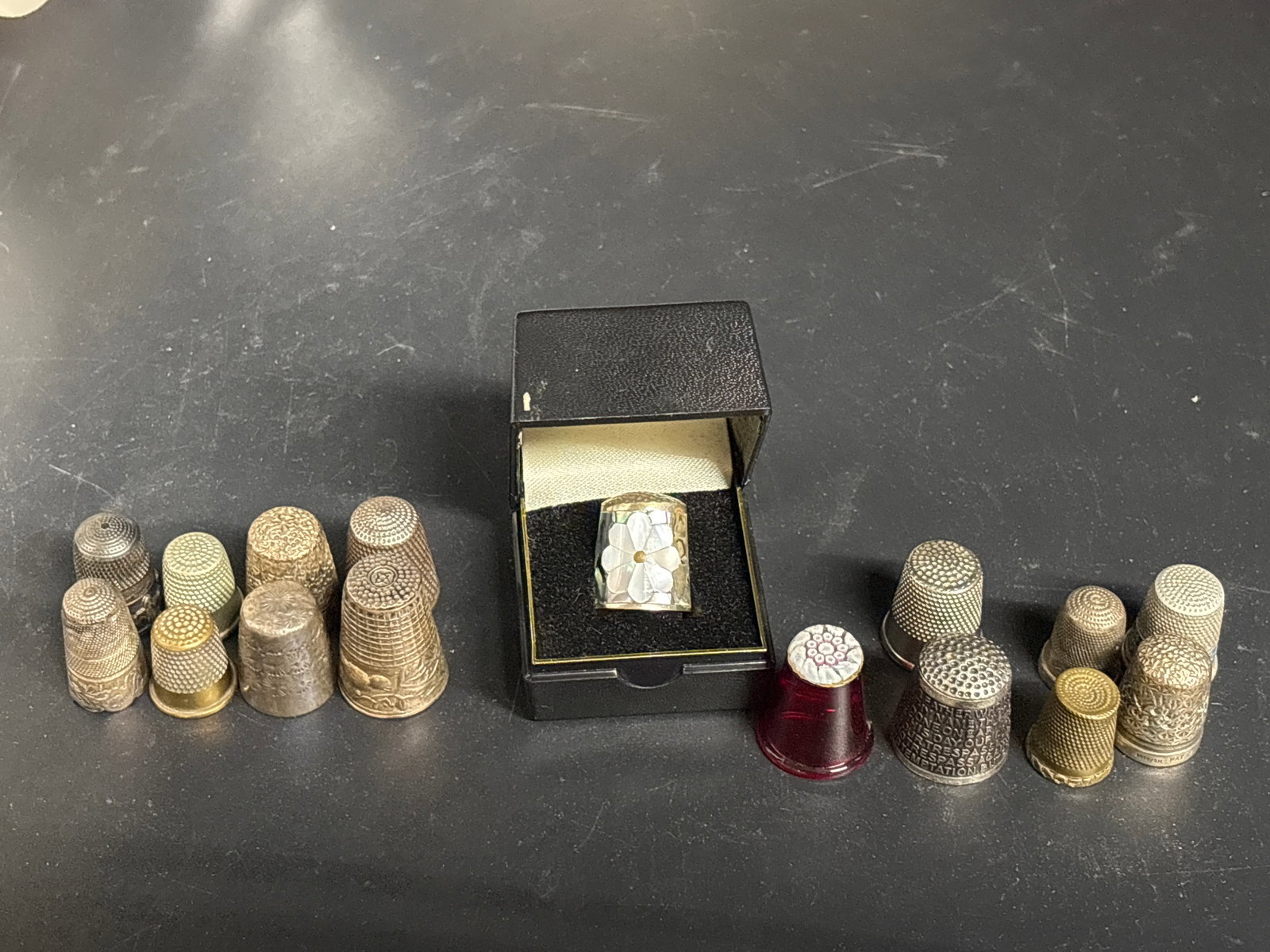 Sixteen assorted antique thimbles; M.O.P., millefiori, Lord’s Prayer, etc.: vintage thimbles.One thimble features a red glass body with a millefiori (thousand flowers) pattern on top, similar to Caithness glass designs.Two other thimbles appear to be standard metallic, textur