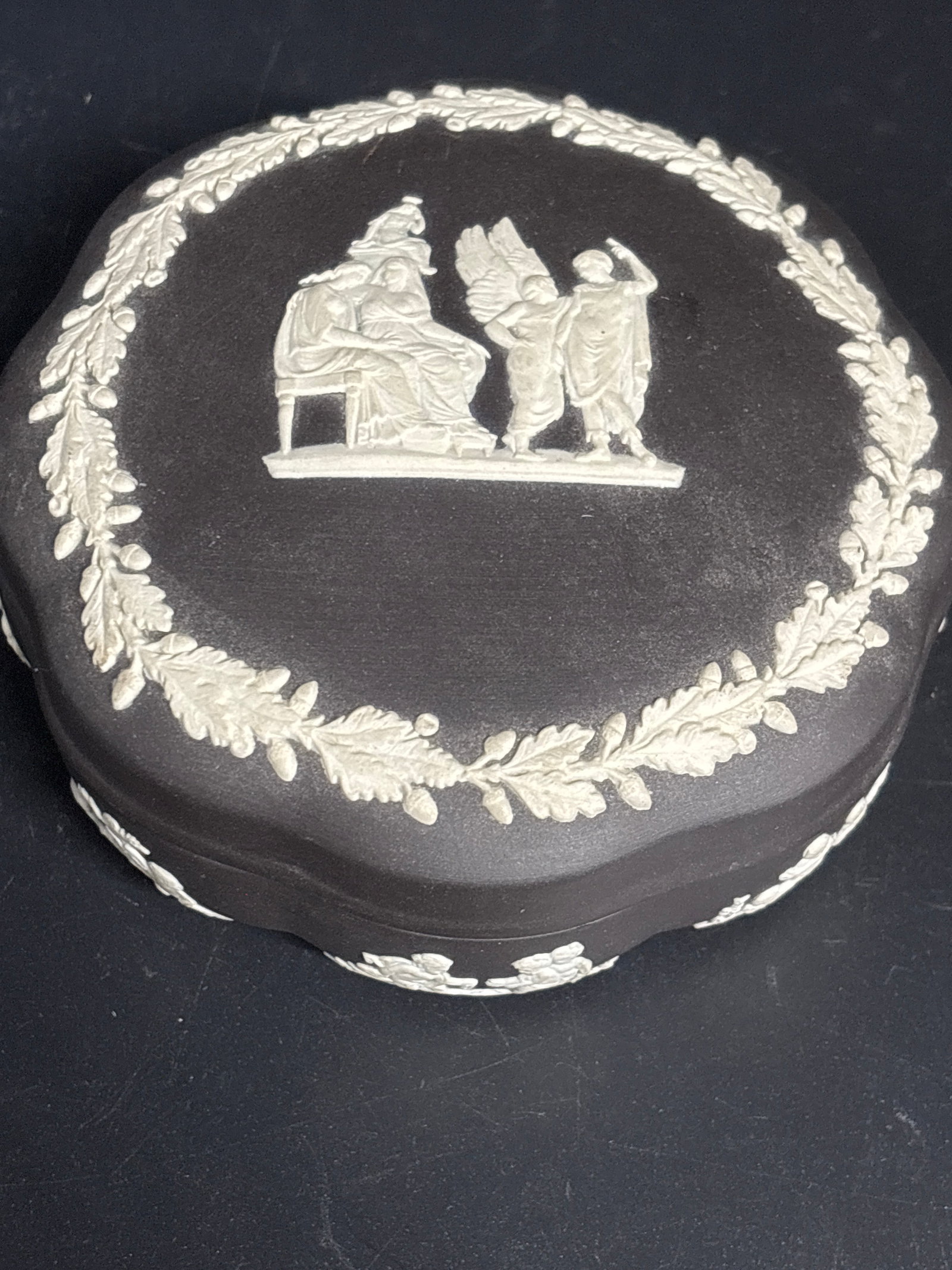 vintage 5 inch Wedgwood black basalt jasperware circular shaped trinket box: vintage Wedgwood Black Jasperware trinket box, crafted in England with a distinctive scalloped edge.The lid features traditional white neoclassical sprigged figures, a style popular in the 1960s and
