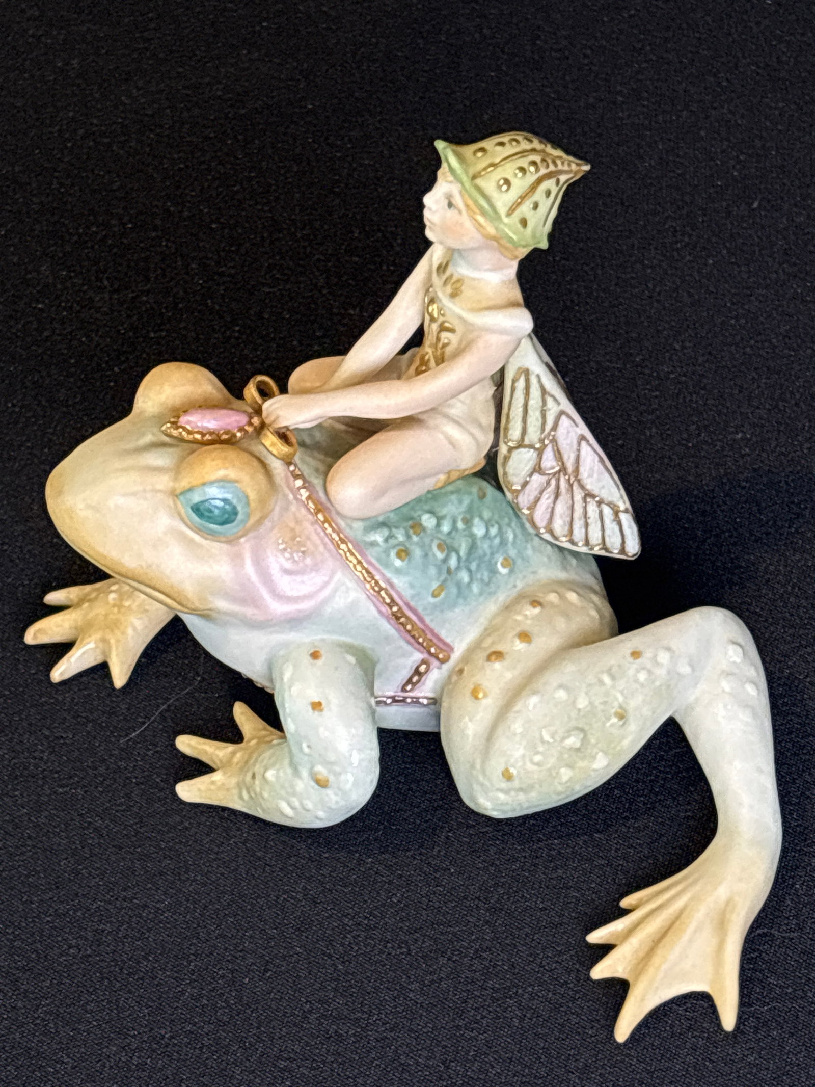 "Tiffin," a winged fairy riding a frog from the "Land of Chimeric" 5 inch figurine: Cybis porcelain figurine titled "Tiffin," a winged fairy riding a frog from the "Land of Chimeric" collection.The piece features a cream-colored frog with textured warts and a fairy with sparkling win