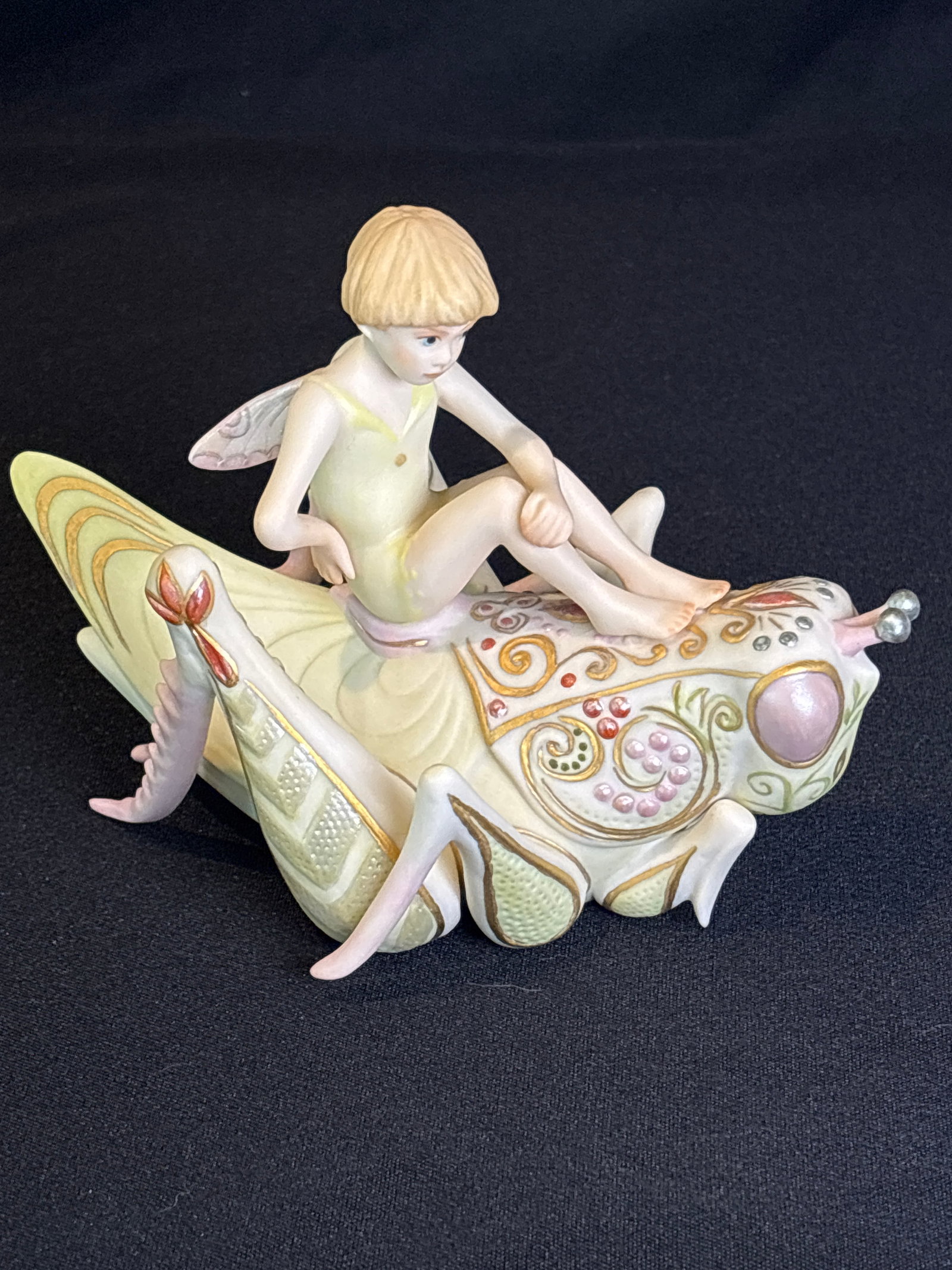 "Ariel" a winged fairy riding a grasshopper from the "Flights of Fancy" 5 inch figurine: Cybis porcelain figurine titled "Ariel," featuring a fairy child riding a grasshopper.Introduced in 1981, this piece was part of the "Flights of Fantasy" collection and was retired in 1983.The figurin