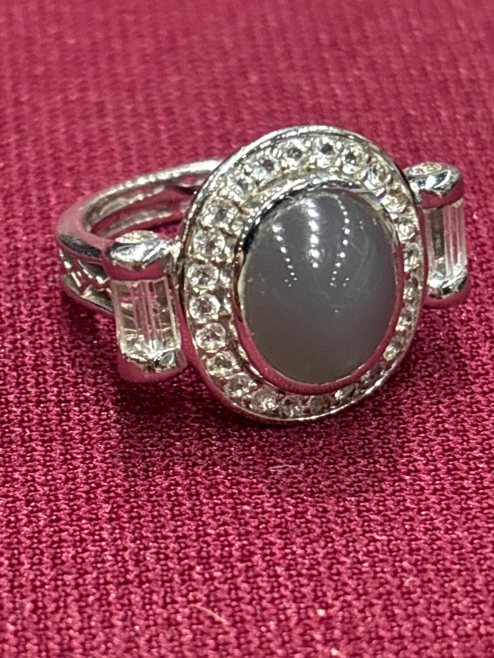 gray cabochon moonstone ring features a halo of smaller stones and baguette side Accents: gray cabochon gemstone ring features a halo of smaller stones and baguette side accents.Stone Type: The center stone is likely a gray moonstone or a gray star sapphire.Material: The setting appears to