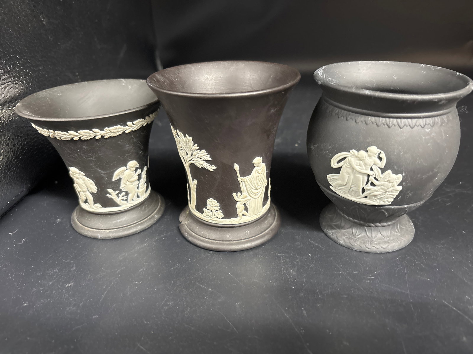 Three similar signed vintage Wedgwood Jasperware black basalt with white relief vases: Three similar signed vintage Wedgwood Jasperware bud vases in black basalt with white classical relief detailing.The vases typically feature cherubs or pastoral scenes inspired by ancient Greek and Ro