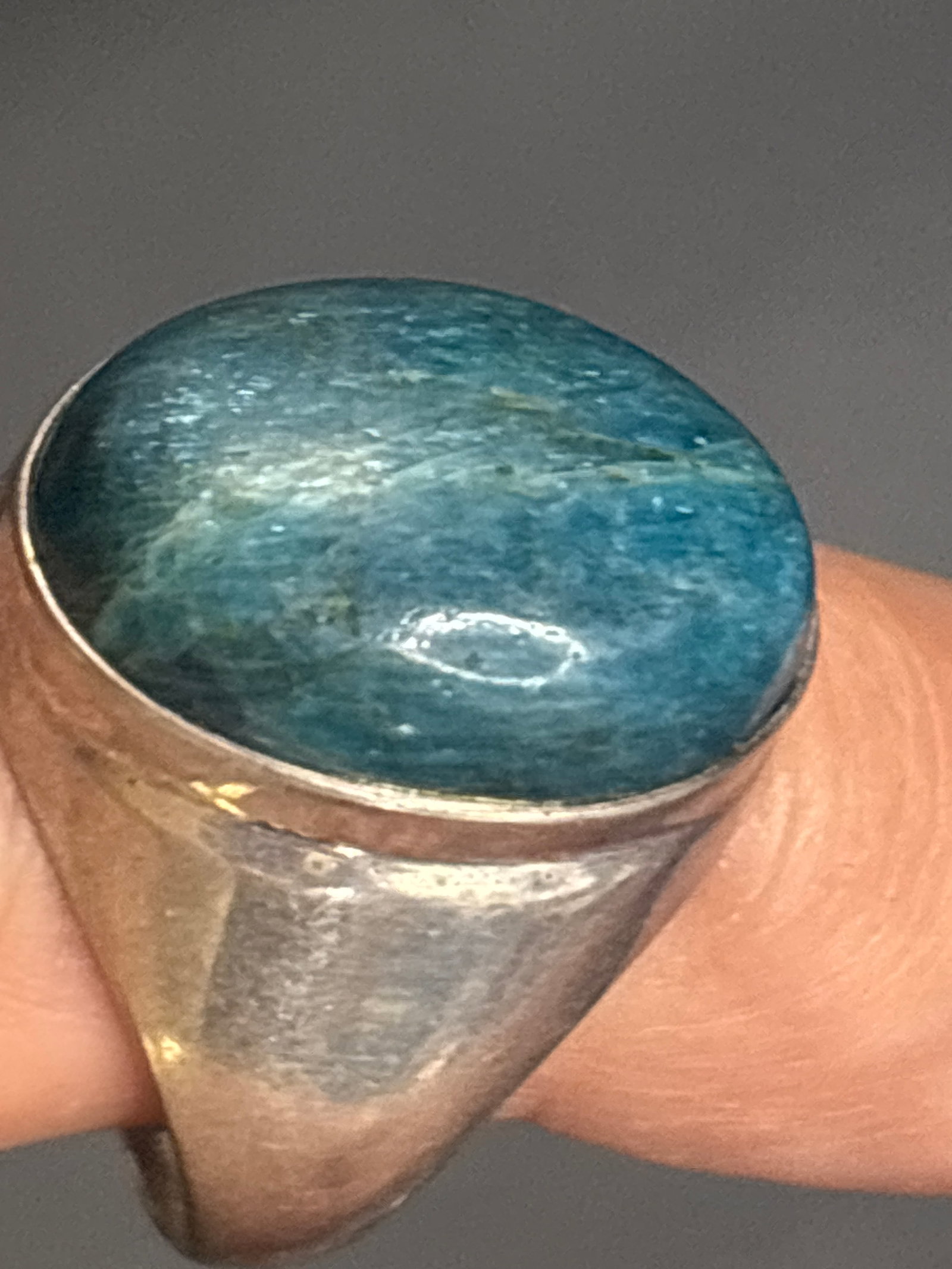 Sterling silver oval, teal-blue cabochon gemstone ring set: Sterling silver oval, teal-blue cabochon gemstone ring set in a chunky silver bezel band. Based on its distinct teal-blue hue, subtle parallel banding, and glassy luster, the stone strongly resembles 