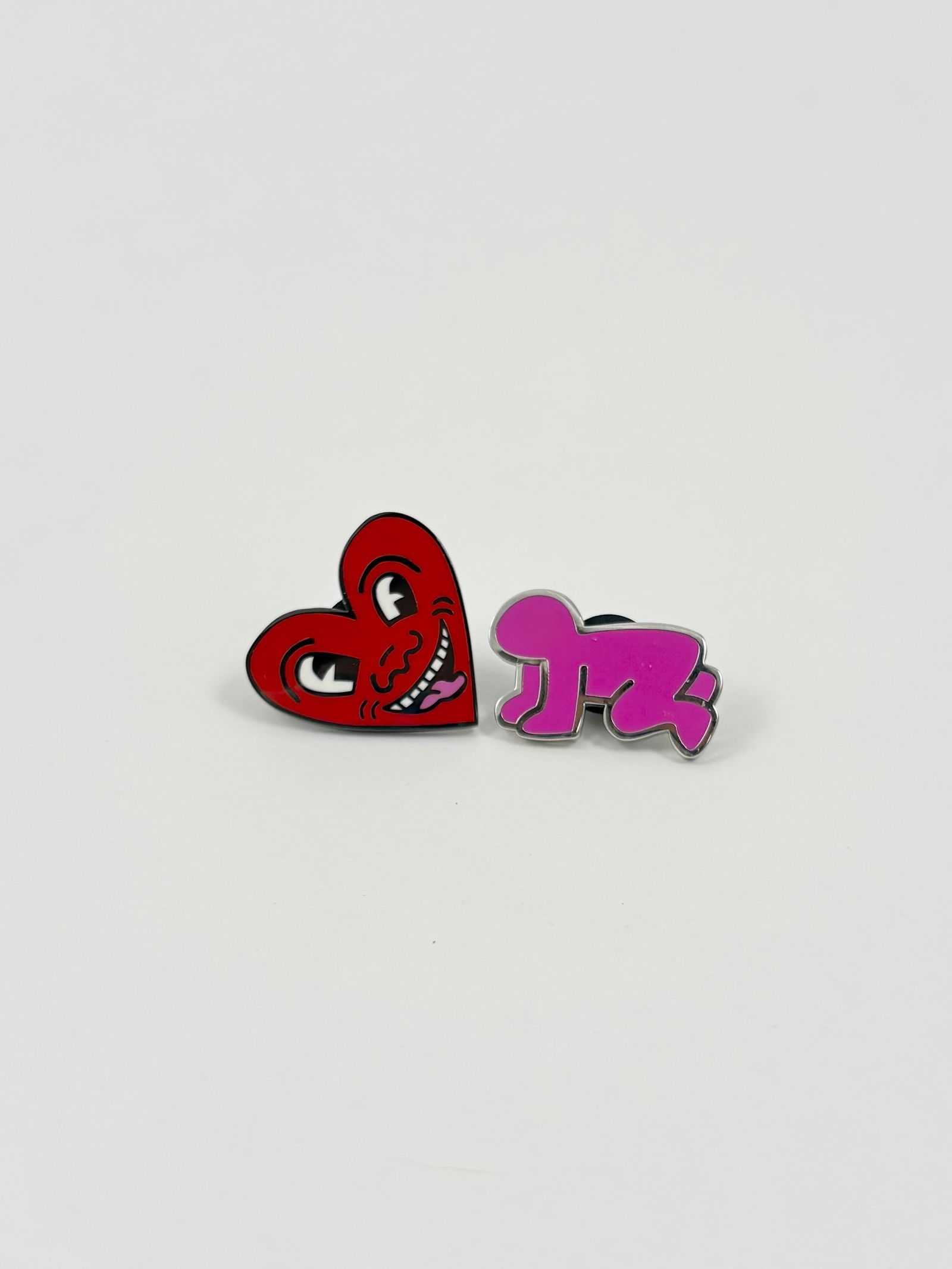 Grouping of Two Keith Haring Enamel Pins: Keith Haring Enamel pins on metal Grouping of two enamel pins after designs by Keith Haring, including a heart motif and a crawling figure form. Both pins feature bold graphic outlines and bright