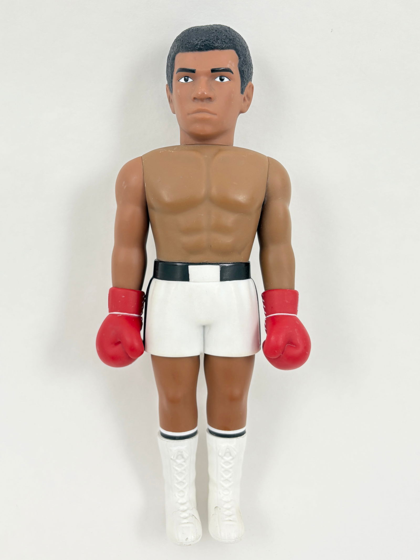 Muhammad Ali - Figure: Collectible figure depicting Muhammad Ali in classic boxing attire, featuring white trunks, red gloves, and lace-up boots. Molded in a standing pose with stylized facial features and athletic