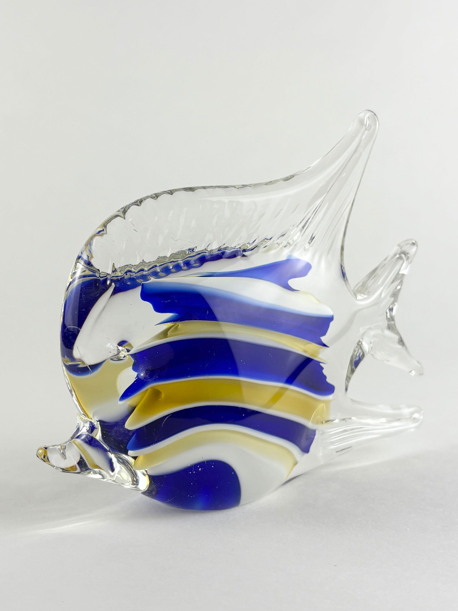Glass Fish Sculpture, Murano Style: Decorative hand-blown glass fish sculpture featuring a rounded body with applied fins and tail. Executed in clear glass with layered blue, white, and yellow ribboned striping throughout the form. Desi