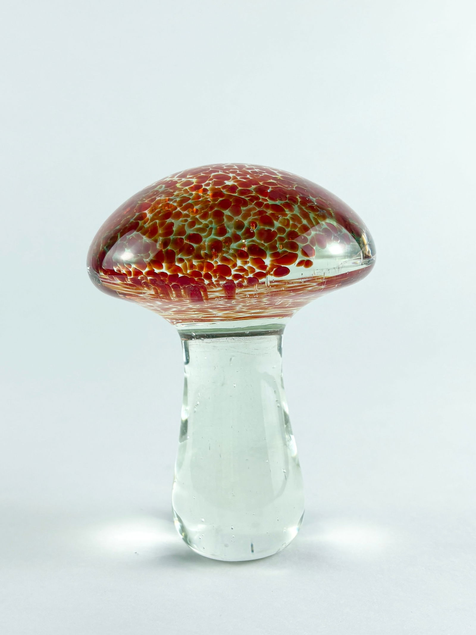 Glass Mushroom Sculpture: Hand-blown glass mushroom sculpture featuring a rounded domed cap in deep red with mottled amber and gold speckling, set atop a clear cylindrical stem. The contrasting textures and colors create a