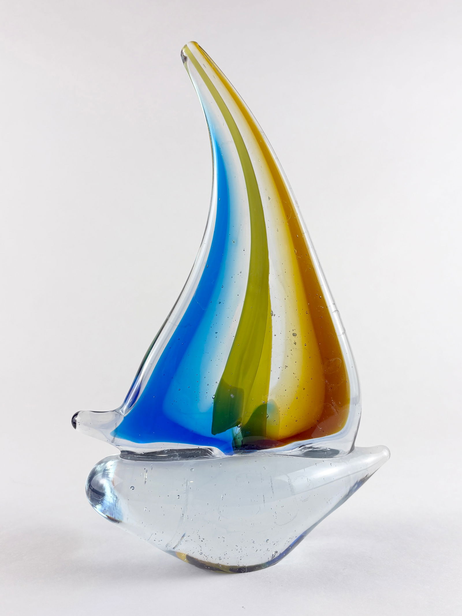 Glass Sailboat Sculpture: Hand-blown glass sailboat sculpture with a clear hull and two elongated sails rendered in layered blue, green, and amber glass. The asymmetrical form and smooth contours give the piece a balanced,