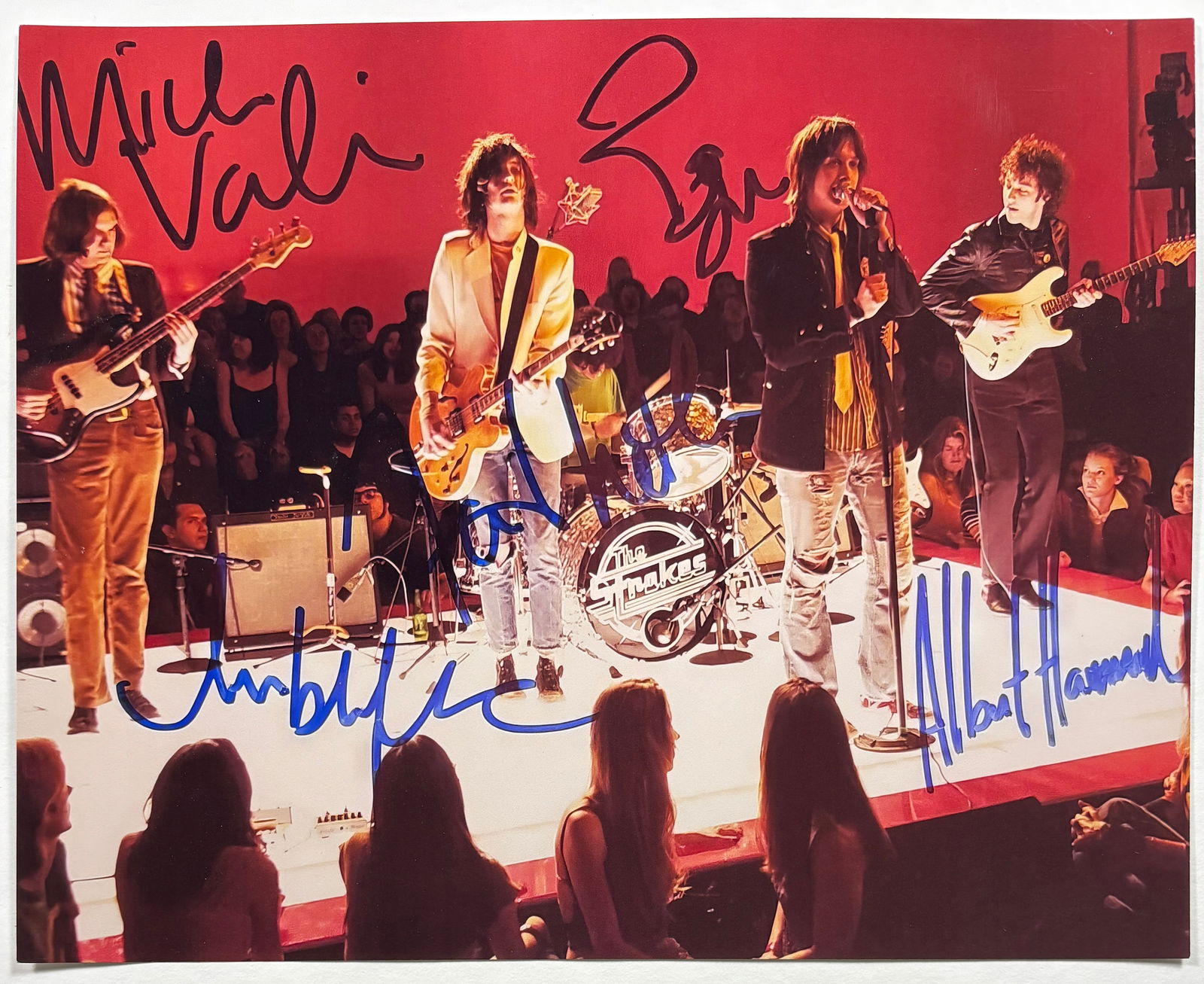 The Strokes Signed, Printed Photo: Printed color photograph depicting the band The Strokes performing live on stage, signed in marker by multiple band members across the image. The photo captures a concert scene with the group
