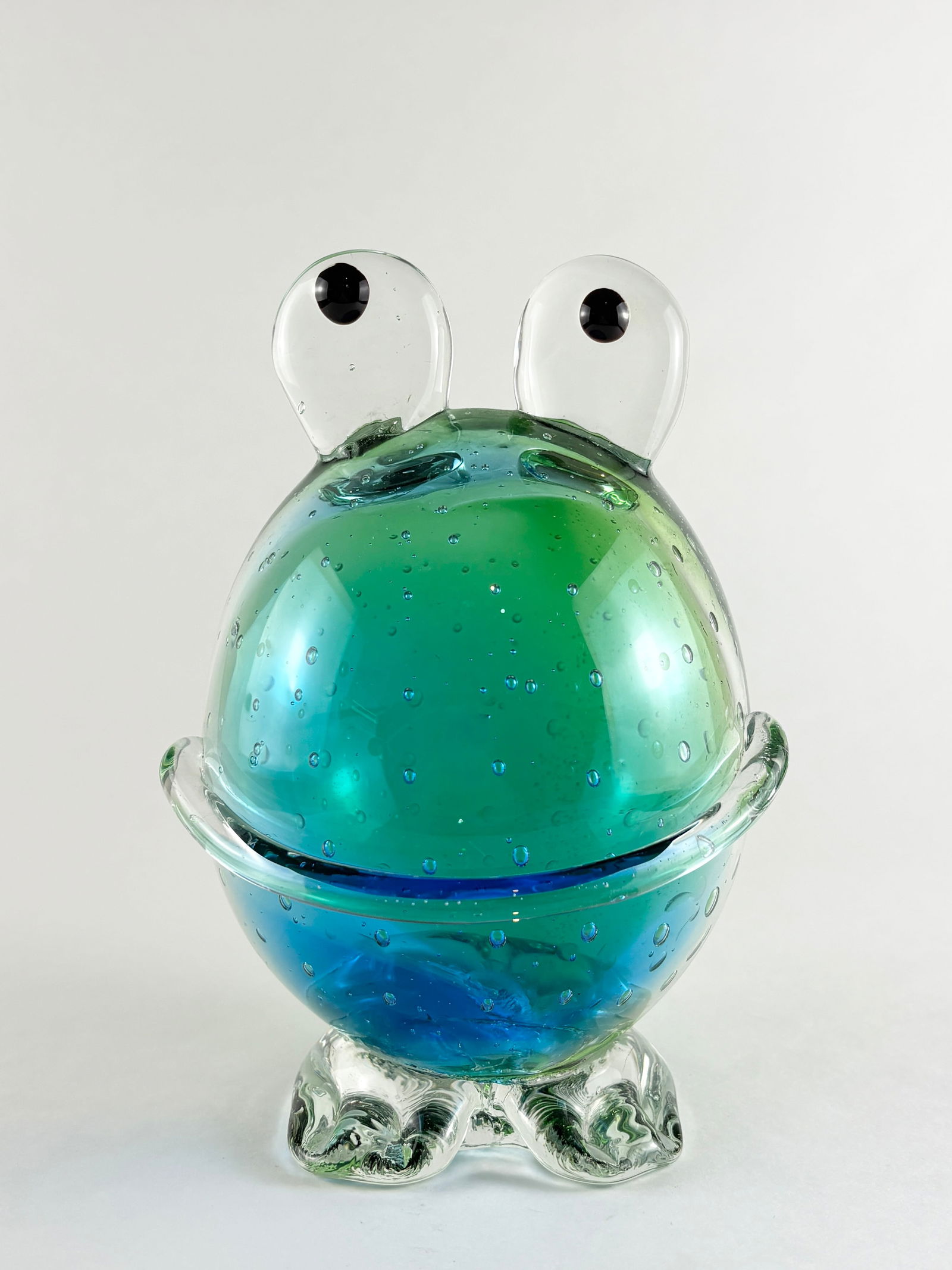 Glass Frog Sculpture: Hand-blown glass frog sculpture with a rounded body form, applied eyes, and integrated feet. Executed in layered green and blue glass with internal bubbles and a smooth polished surface. Decorative