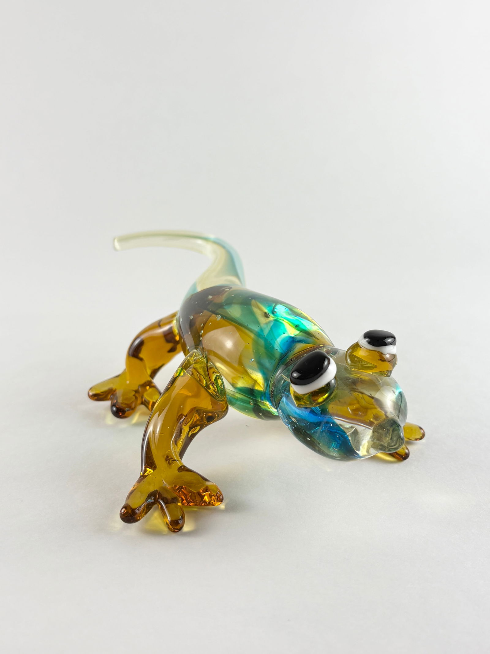 Murano Style Glass Lizard Sculpture: Murano style hand-blown glass lizard sculpture executed in translucent amber and aqua tones, with applied eyes and elongated tail forming a dynamic, low-profile pose. The piece shows decorative surfac