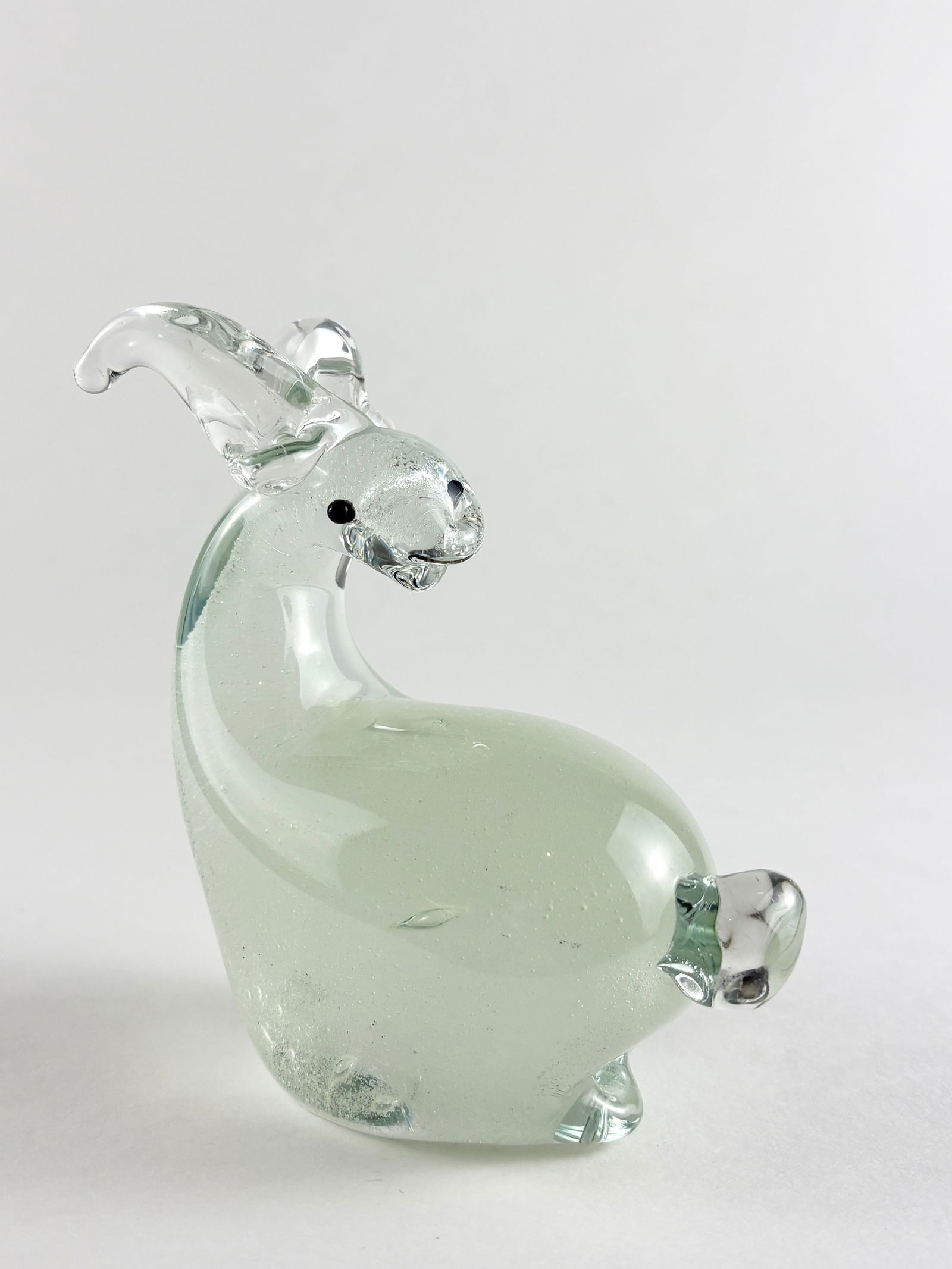 Glass Goat Sculpture: Decorative clear and pale green glass goat sculpture with a rounded body, raised head, and applied horn and leg details. The piece features a smooth, translucent finish with simple sculptural modeling