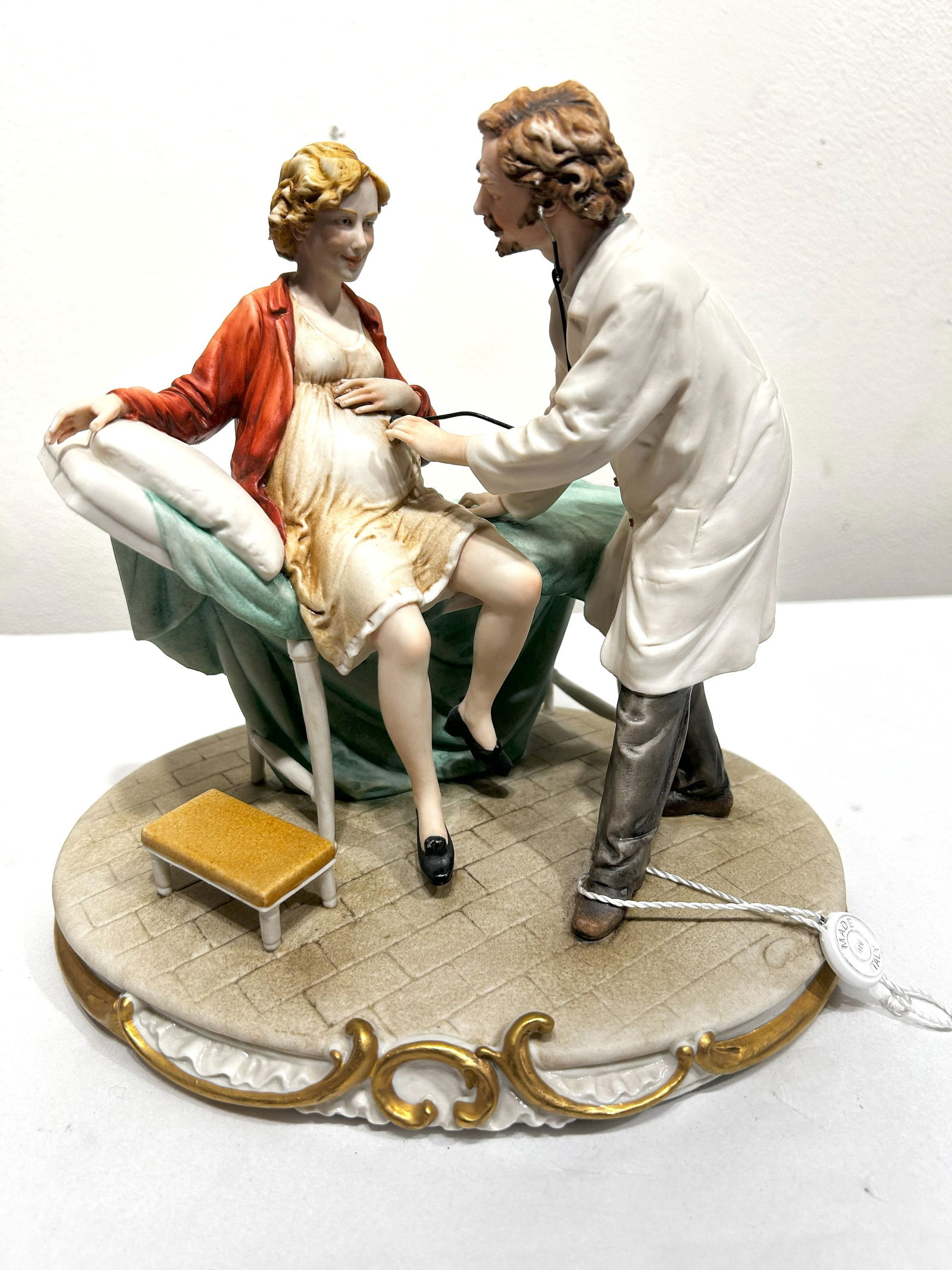 Capodimonte Porcelain Figurine: Doctor Examining Pregnant Woman: This detailed Capodimonte porcelain figurine depicts a tender scene of a doctor examining a pregnant woman. The doctor, dressed in a white coat, is using a stethoscope to listen to the expectant mothe