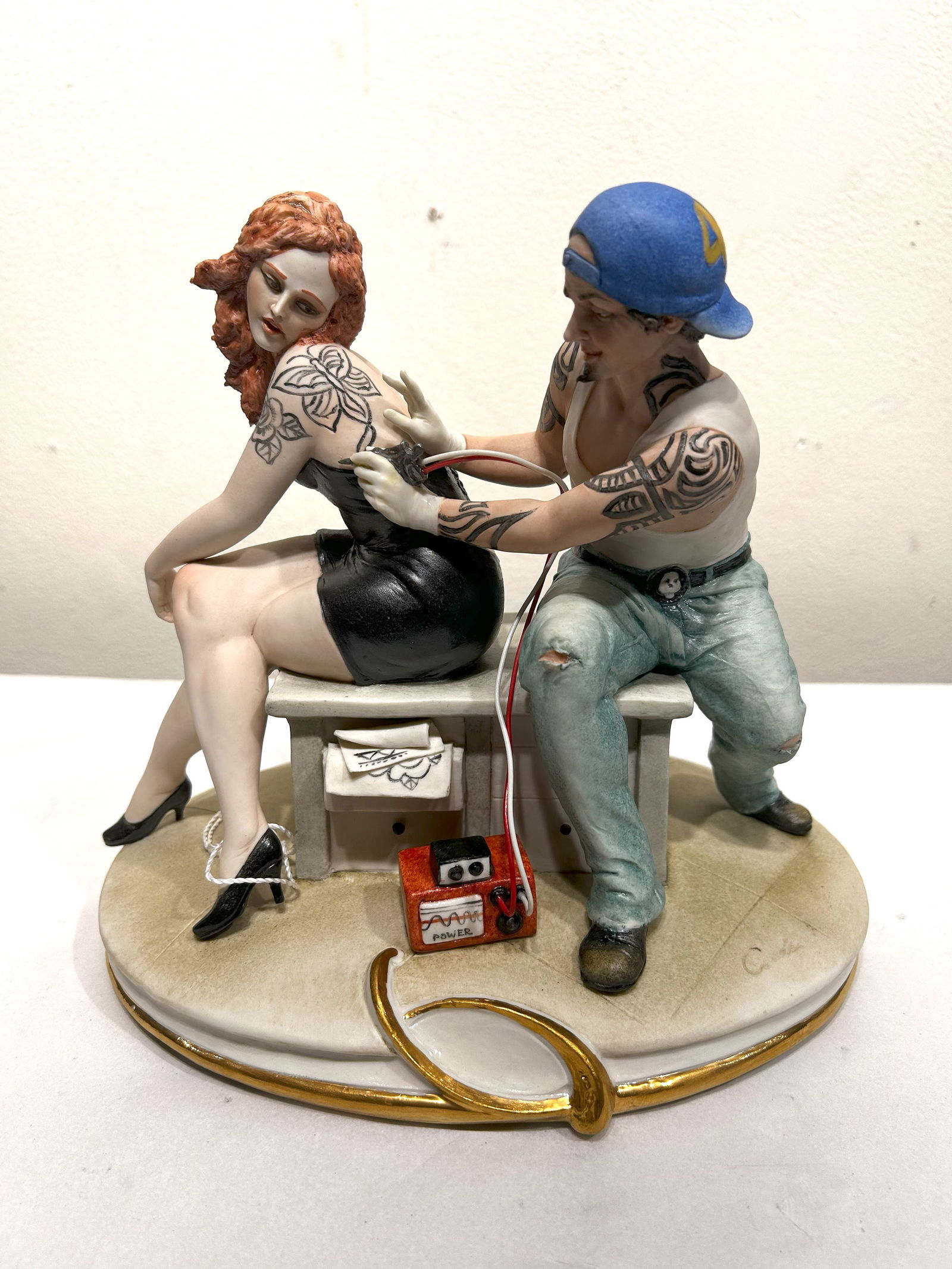 Capodimonte Porcelain Figurine: Tattoo Artist at Work: This detailed Capodimonte porcelain figurine captures a scene of a tattoo artist inking a design onto a woman's back. The artist, wearing a blue baseball cap and jeans, is carefully working on the sea