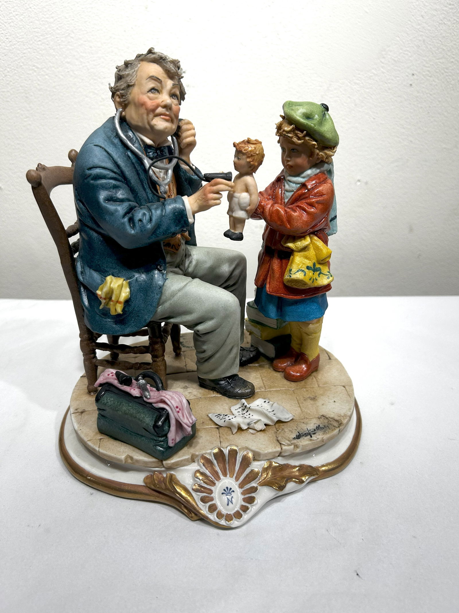 Capodimonte Porcelain Figurine: Doctor Examining Child with Doll: This charming Capodimonte porcelain figurine depicts a doctor in a blue coat, seated and examining a child holding a doll. The child, wearing a green beret and a red coat, stands next to the doctor, p