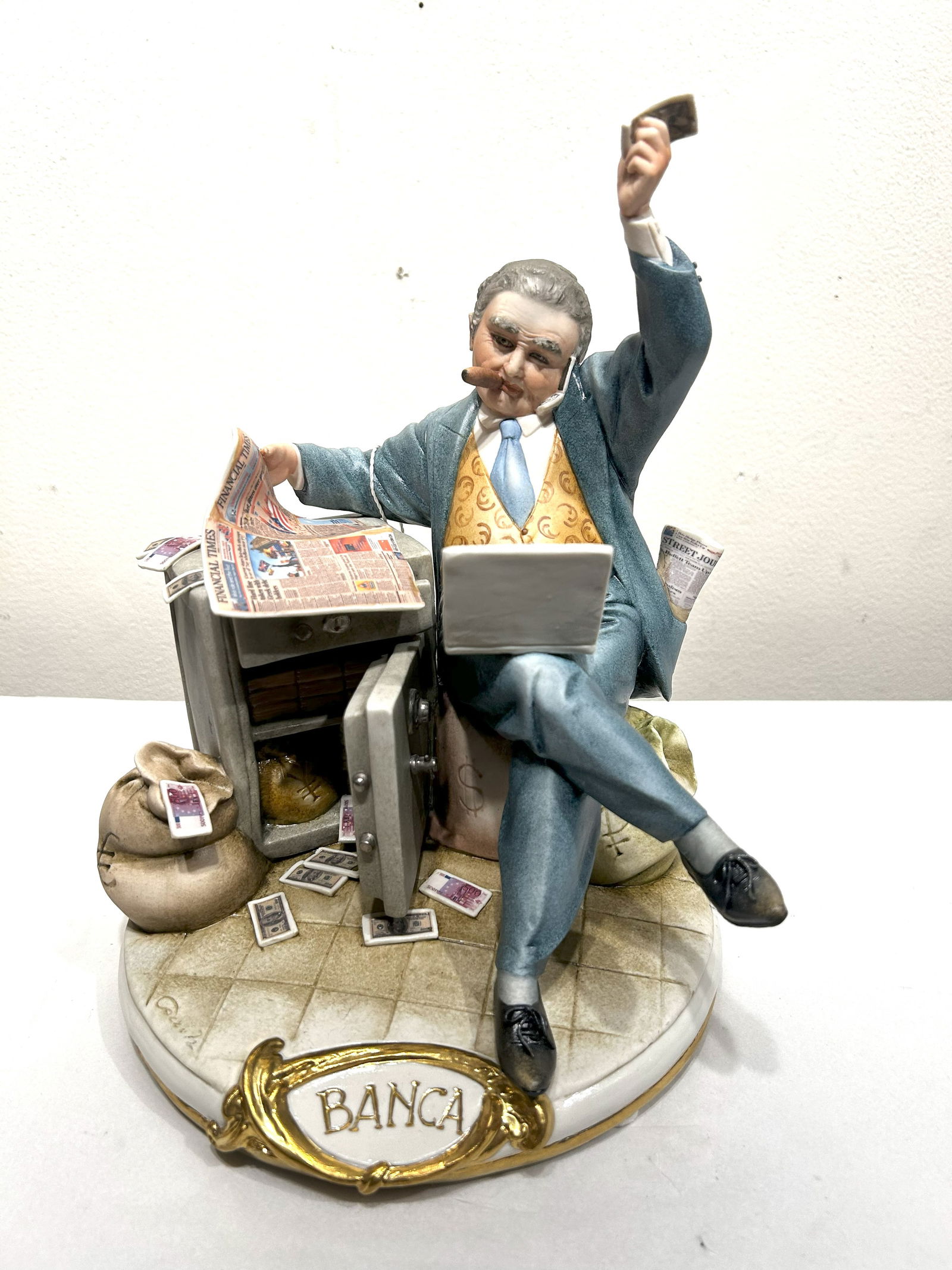 Capodimonte Porcelain Figurine: The Banker: This detailed Capodimonte porcelain figurine depicts a banker sitting amidst symbols of wealth. He's shown with a cigar, a laptop, and surrounded by open safes overflowing with money, bags of currency