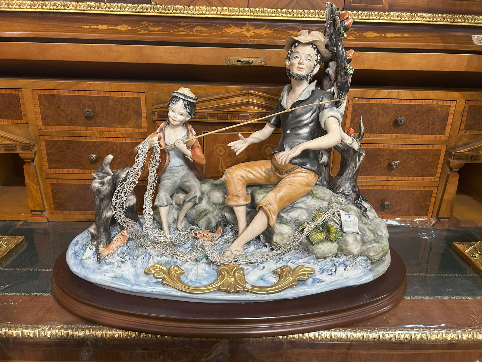 Large Capodimonte Sartori Fisherman Figurine: This impressive Capodimonte porcelain figurine, signed by Sartori, depicts a detailed scene of a fisherman and a young boy with their catch. The fisherman is seated on rocks, holding a fishing rod, wh