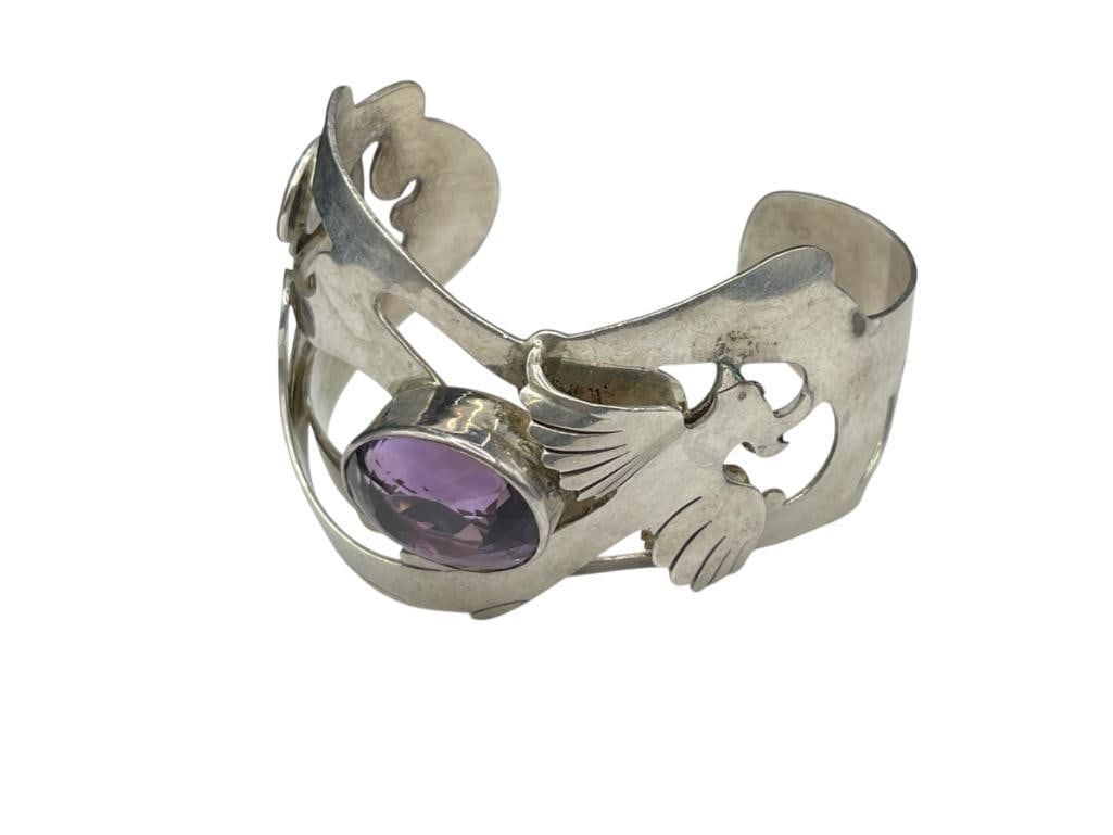 VINTAGE ARTIST SIGNED STERLING SILVER BIRD AND AMETHYST CUFF BRACELET: FEATURING BIRD FLYING ABOVE AN OVAL CUT AMETHYST 2 1/2" CUFF WIDTH, 1 5/8" FRONT, 20MM X 16MM AMETHYST, 49.27 GRAMS 