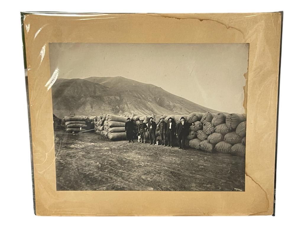 1920'S BLACK AND WHITE PHOTOGRAPH OF WOOL GROWERS IN NEVADA: PHOTO DEPICTS SIX MEN WITH DOGS STANDING IN FRONT OF LARGE SACKS FILLED WITH WOOL, LIGHT WATER DAMAGE RIGHT EDGE 9 1/2" X 7 1/4" 