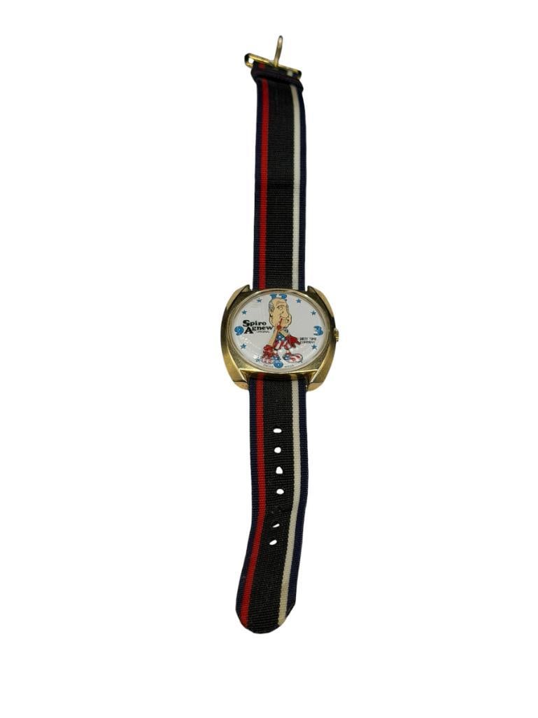 VINTAGE SPIRO AGNEW DIRTY TIME COMPANY WRISTWATCH FEATURING CARTOON IMAGE OF U.S. VICE PRESIDENT: FIGURAL HANDS FOR WATCH HANDS, NYLON BAND, CLASP MARKED HONG KONG, SWISS MADE MOVEMENT, IN RUNNING CONDITION 32.5MM CASE