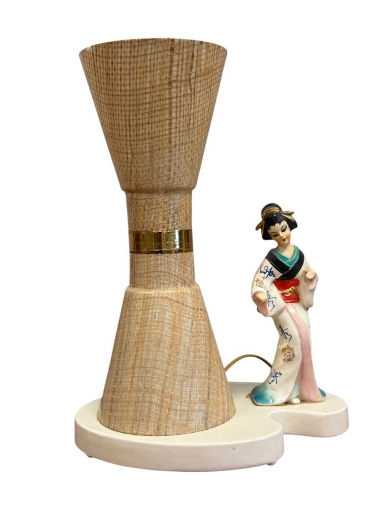 THE VIGON CO. MID-CENTURY FIGURAL LAMP WITH GEISHA FIGURE AND HOUR GLASS SHAPED SHADE LOS ANGELES: LAMP FEATURES A GEISHA FIGURE WHICH HAS BEEN PREVIOUSLY REPAIRED AT THE BASE, WICKER LIKE SHADE HAS CHIPS ALONG THE TOP EDGE, TESTED + 13 1/4" H