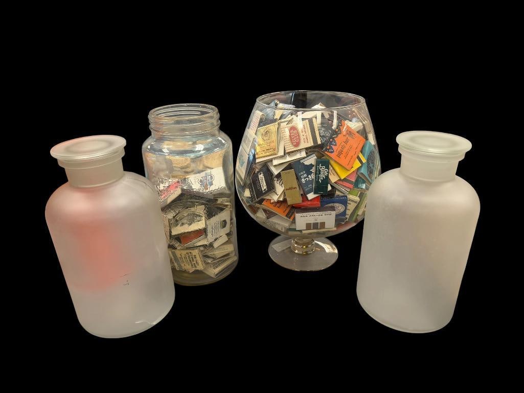 FOUR GLASS CONTAINERS TWO FILLED WITH MANY ASSORTED MATCHBOOKS: INCLUDES MATCHBOOKS FROM HOTELS, CASINOS, RESTAURANTS, ETC., ONE OF THE FROSTED GLASS JARS HAS A CRACK ALONG THE BOTTOM 10", 10 1/2", AND 11" H