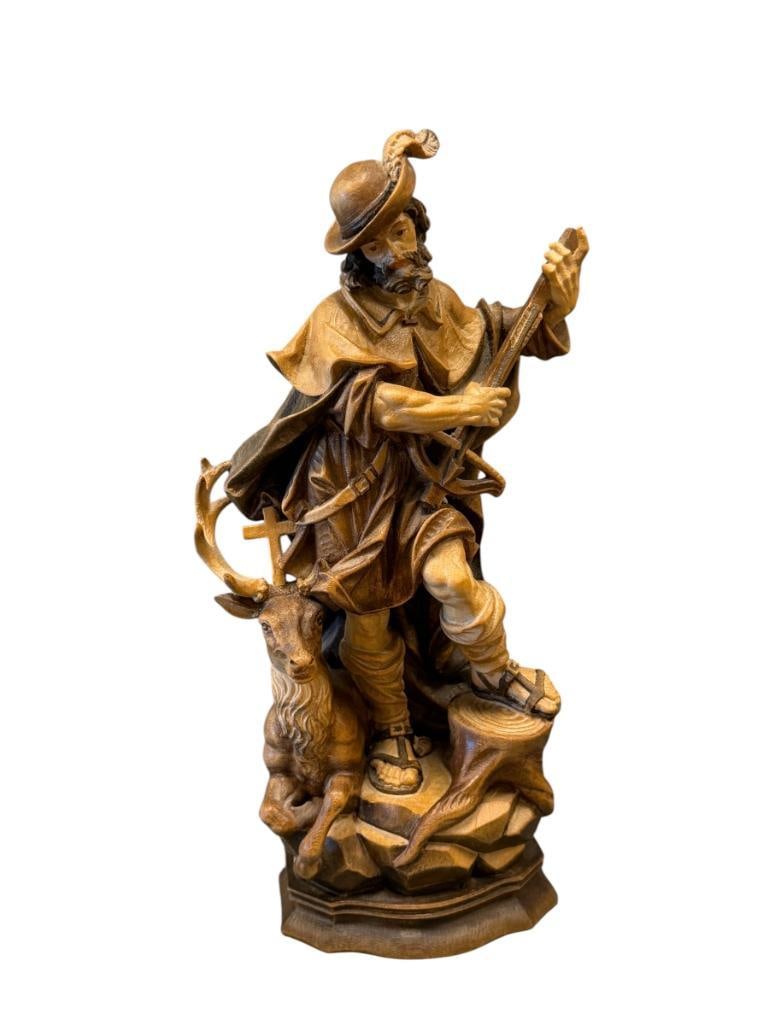 CARVED WOOD SCULPTURE OF SAINT HUNERTUS THE PATRON SAINT OF HUNTERS: TRADITIONAL ALPINE STYLE FEATURES MULTIPLE TONES, DEPICTS SAINT IN HUNTING ATTIRE HOLDING A BOW WITH STAG ALONG THE BASE 11 5/8" H
