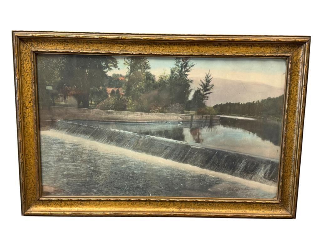 EARLY 20TH CENTURY HAND COLORED PHOTOGRAPH OF A SCENE OF THE TRUCKEE RIVER: PHOTO DEPICTS SCENE ON THE TRUCKEE WITH BUILDINGS HIDING BEHIND THICK TREE LINE AND MOUNTAINS IN THE BACKGROUND, FRAMED BY BRUNDIDGES 20" X 13" FRAME