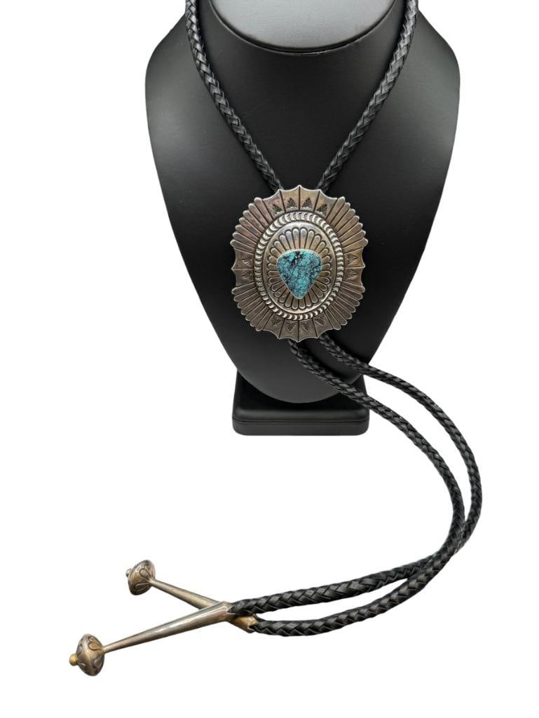NAVAJO JOHN LA RON BEDONI SR ARTIST SIGNED STERLING SILVER TURQUOISE BOLO TIE: FEATURING STAMPED DESIGN AND INTRICATE DETAIL, TRIANGULAR SHAPED TURQUOISE IN CENTER, SIGNED "JB SR NAVAJO", BLACK LEATHER BOLO TIE WITH STERLING SILVER TIPS 3" BOLO CLIP, APPROX. 50 GRAM CLIP, 48"