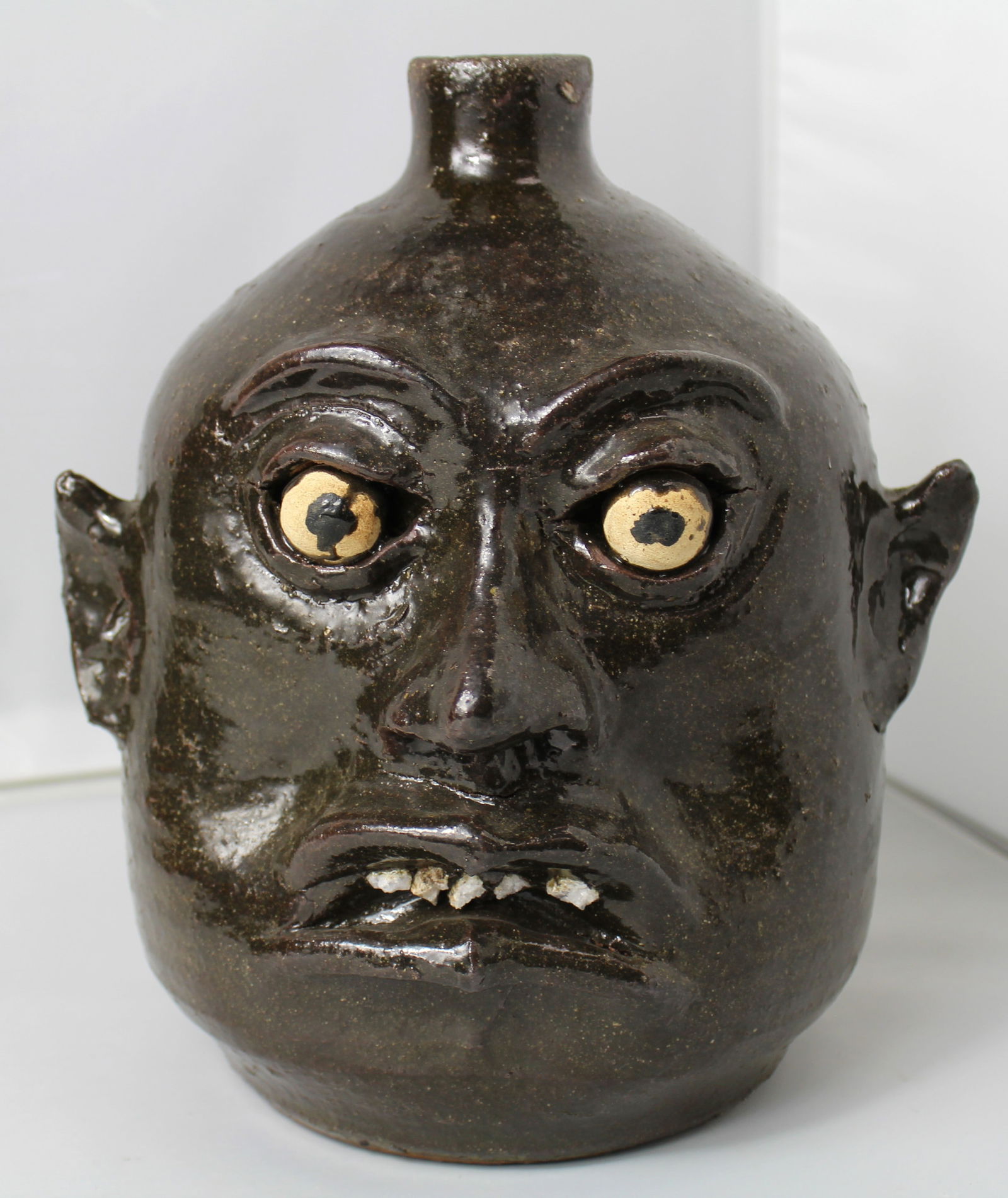 Lanier Meaders (1917-1998) Grotesque Face Jug: Lanier Meaders (1917-1998) Grotesque Face Jug. Unique dark green glazed pottery of a jug with a face with tan eyes and white teeth. The jug is signed on the bottom by the artist. There is a small chip