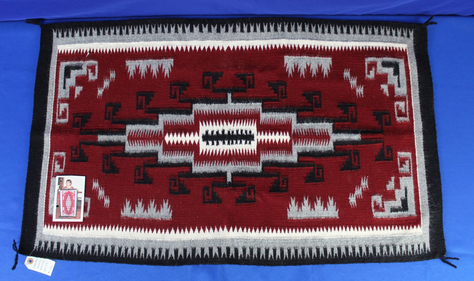 Ganado Navajo Saddle Blanket 1: Red, white, and black Navajo saddle blanket with square and triangle designs. The blanket has four tails on each corner and has a price tag and photo tag of the maker, Annie White. The saddle blanket