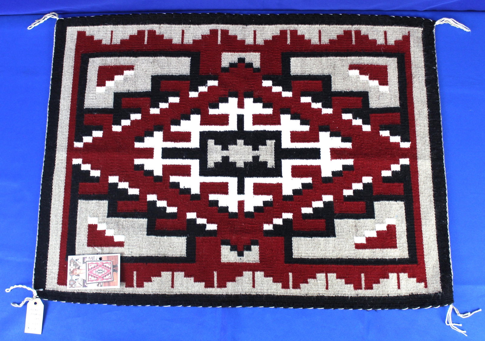 Ganado Navajo Saddle Blanket 2: Red, white, and black Navajo saddle blanket with square and diamond designs. The blanket has four tails on each corner and has a price tag and a photo tag of the maker of the blanket, Lorraine