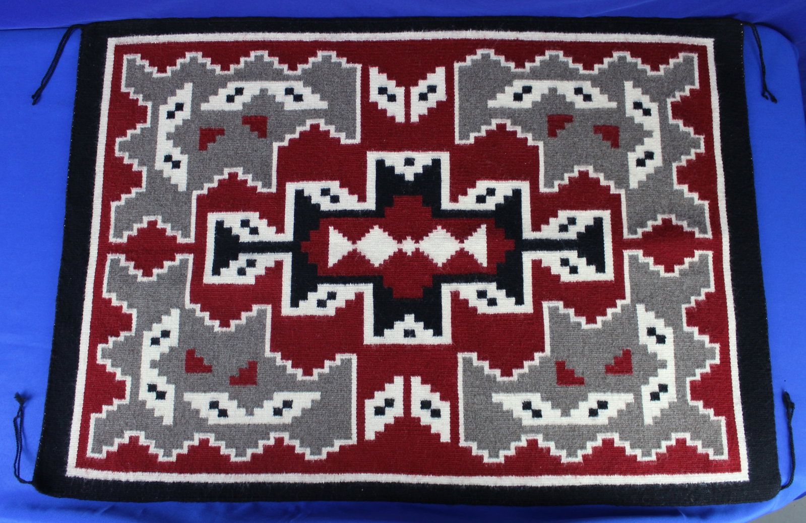 Navajo Saddle Blanket 3: Red, white, and black Navajo saddle blanket with unique square designs. The blanket has four tails on each corner. One of the tails has some light fraying, but overall the saddle blanket is in